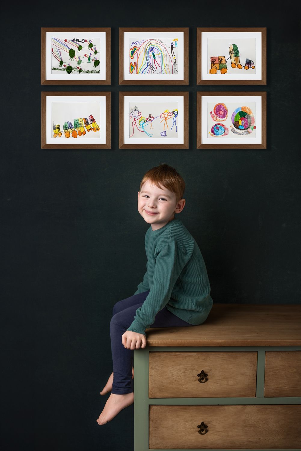 Child in green sweater sitting on wooden dresser with framed artwork displayed above.