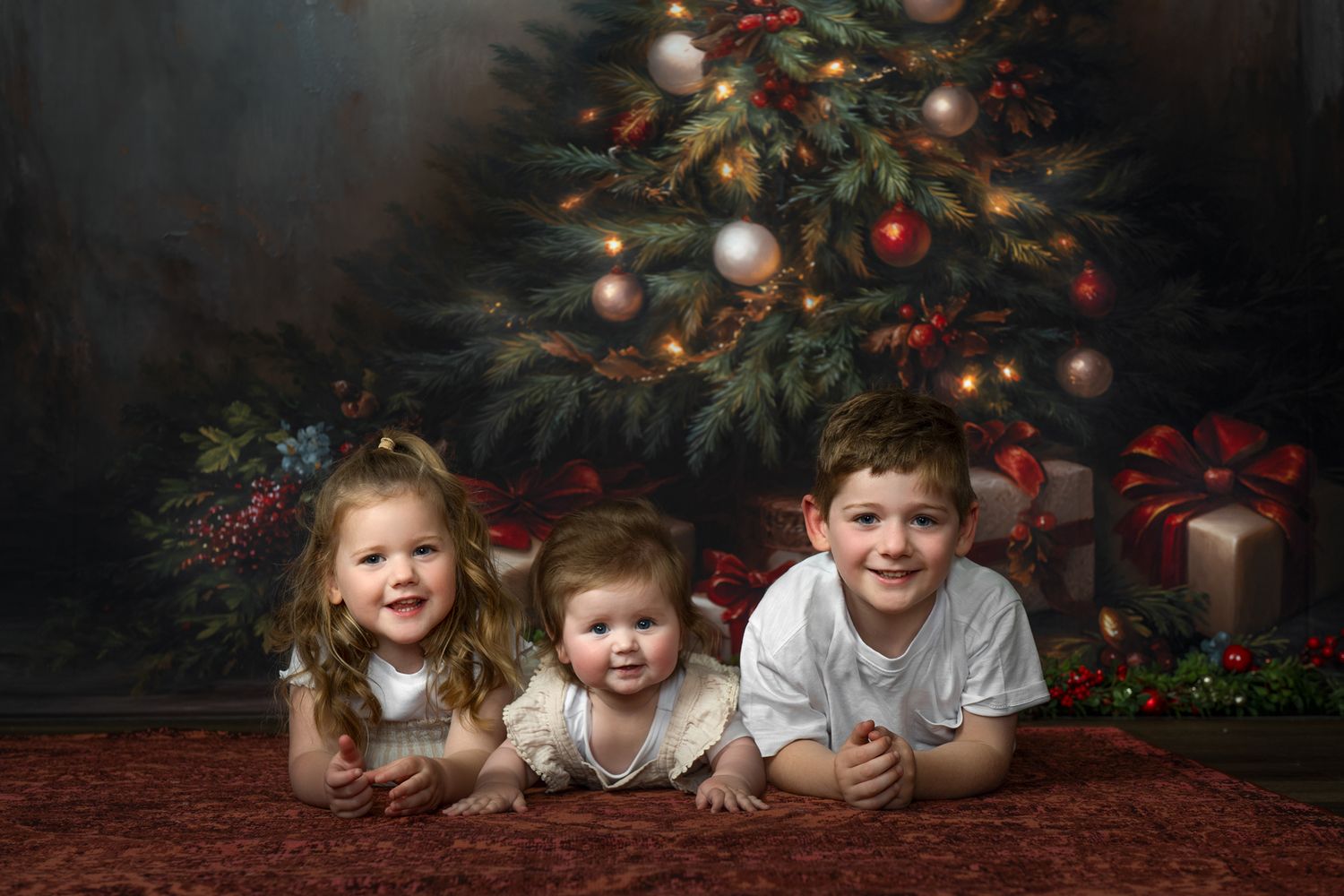 Three children lying on a red carpet in front of a decorated Christmas tree with presents underneath.