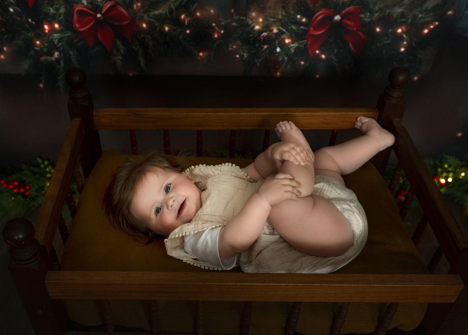 Baby in white dress lying on wooden bench with Christmas lights in background.
