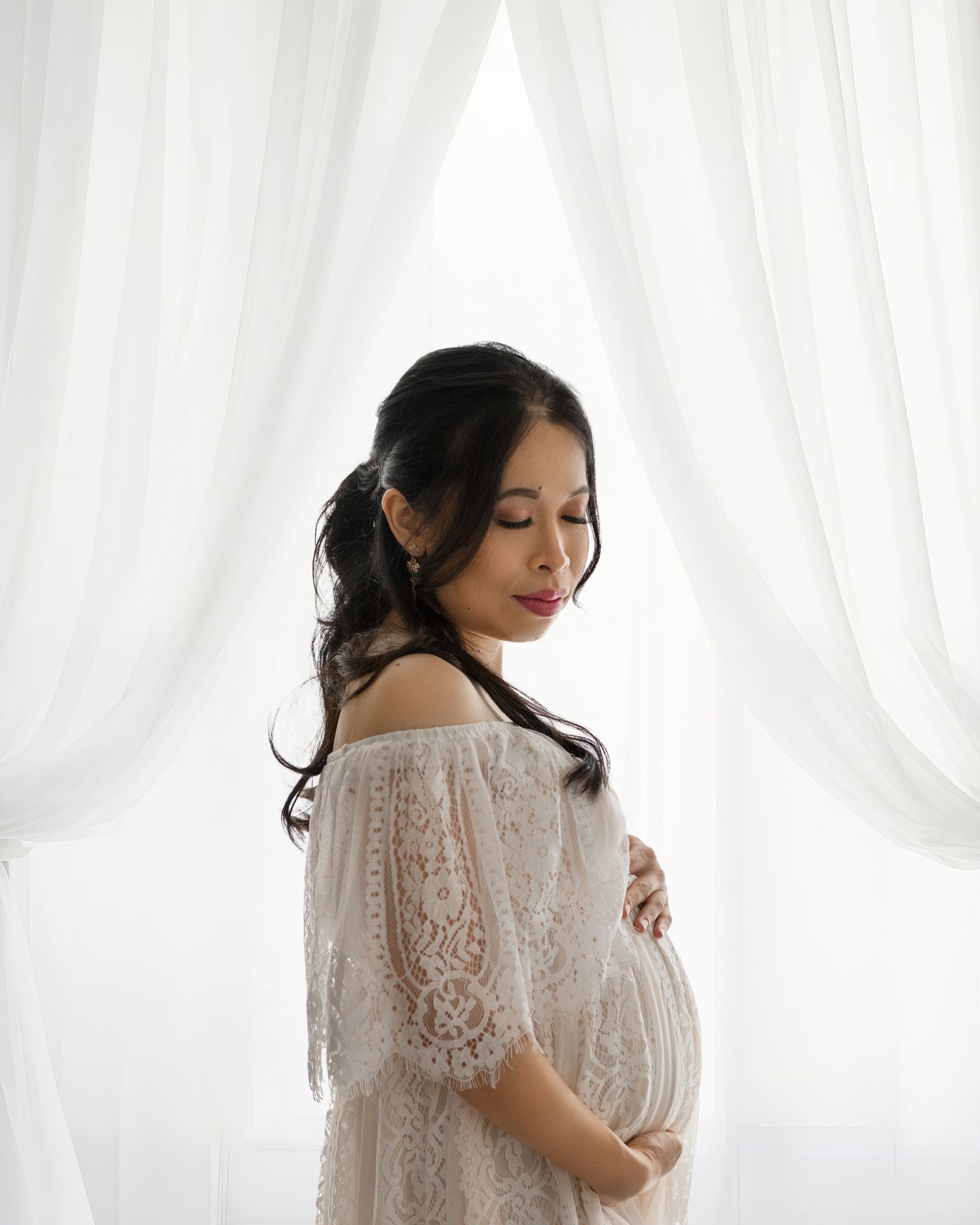 Expectant mother in delicate white lace dress poses against sheer curtains in a dreamy maternity photo.