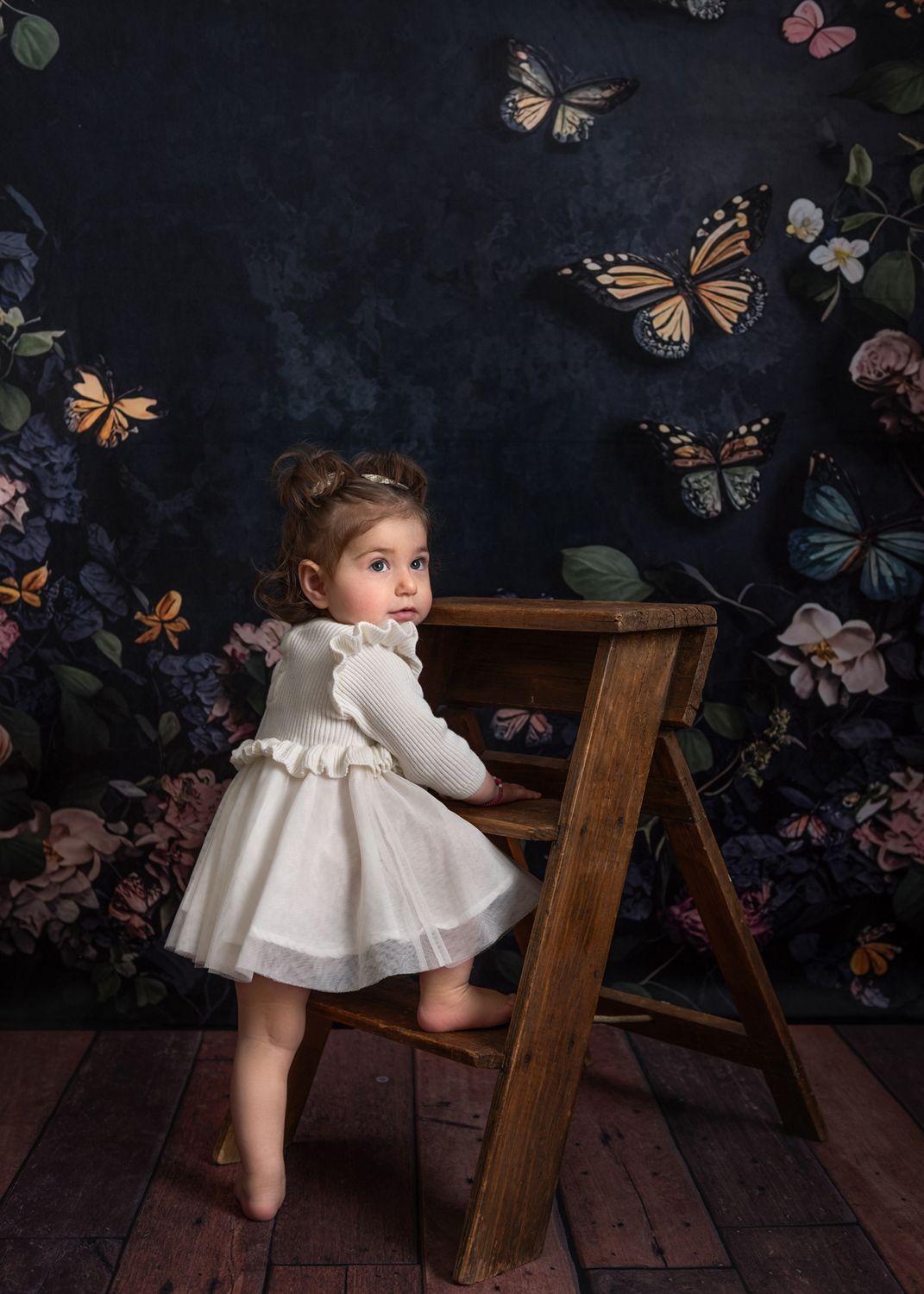 Whimsical baby photo with butterflies and wooden ladder against dark backdrop.