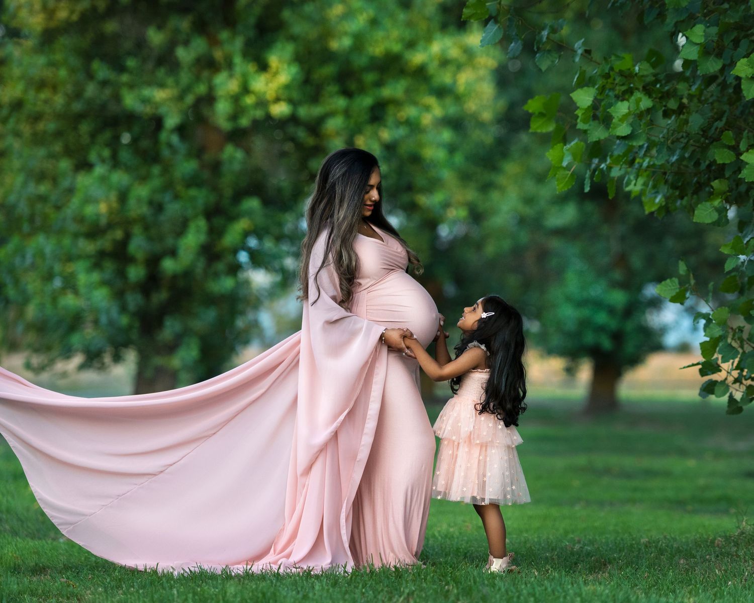 Flowing pink dress billows gracefully across a lush green lawn in an ethereal maternity photoshoot.