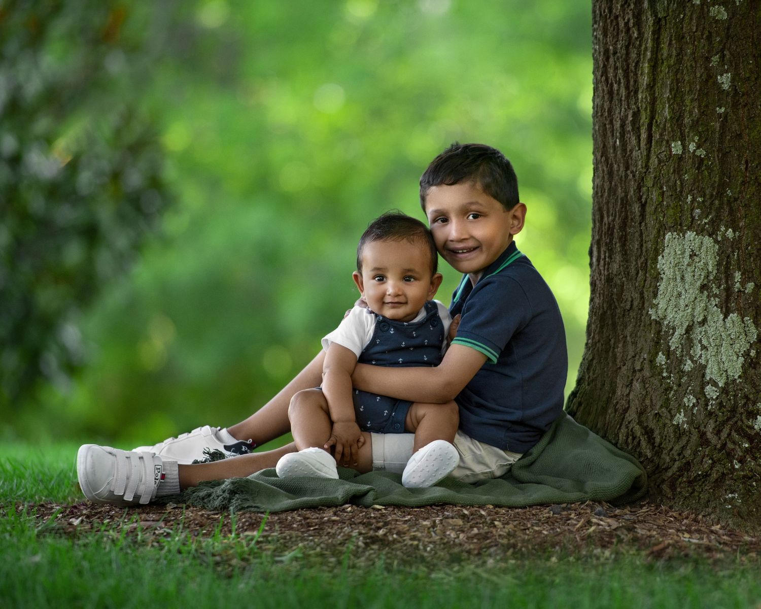 Two children pose together under a tree in casual clothing.