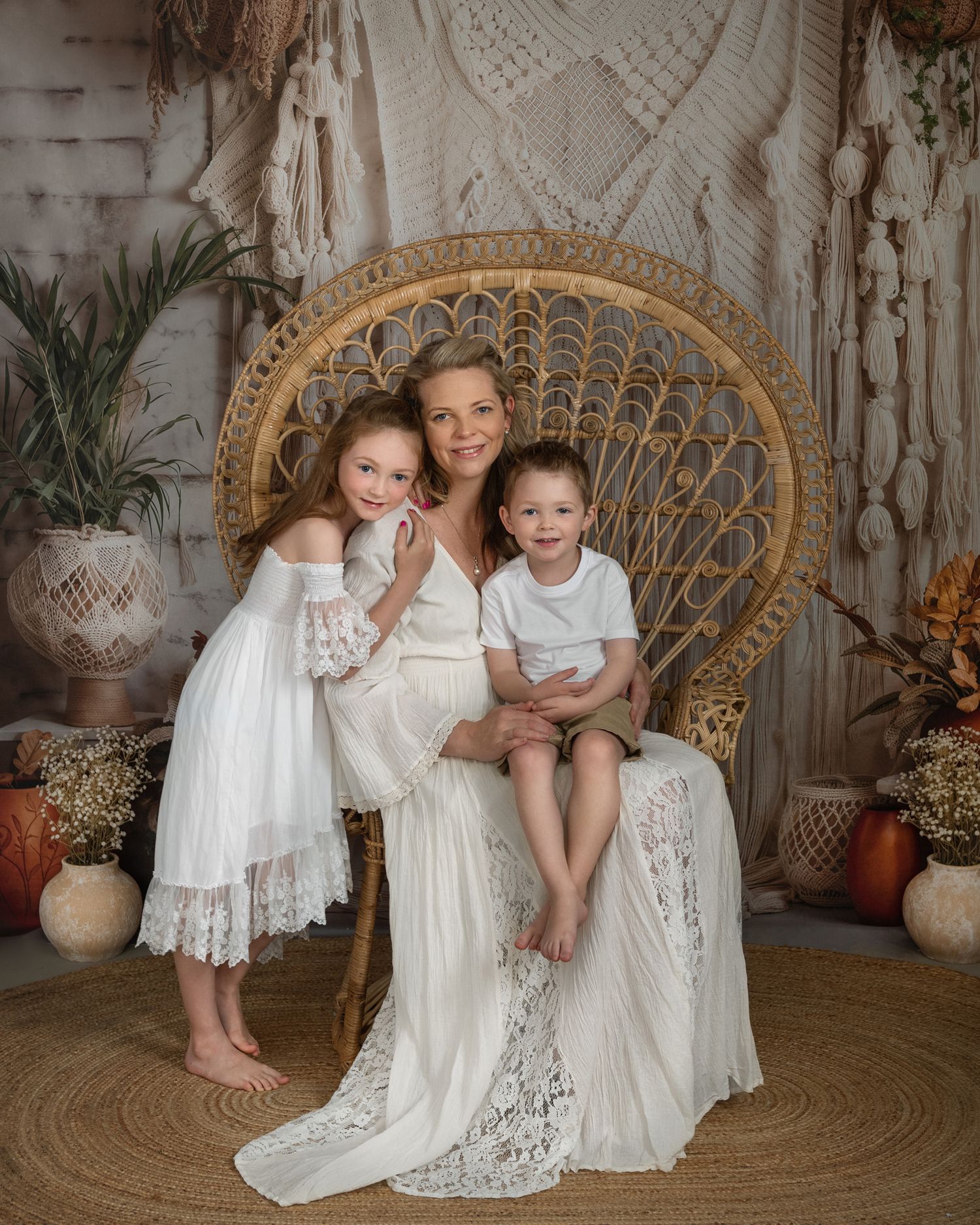 Boho family photoshoot featuring white lace dresses and wicker peacock chair against textured backdrop.
