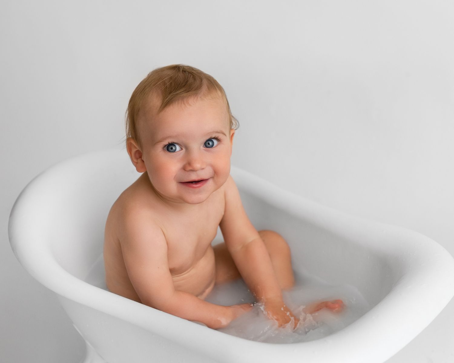 Baby sitting and playing in white porcelain bathtub during milestone photo session.