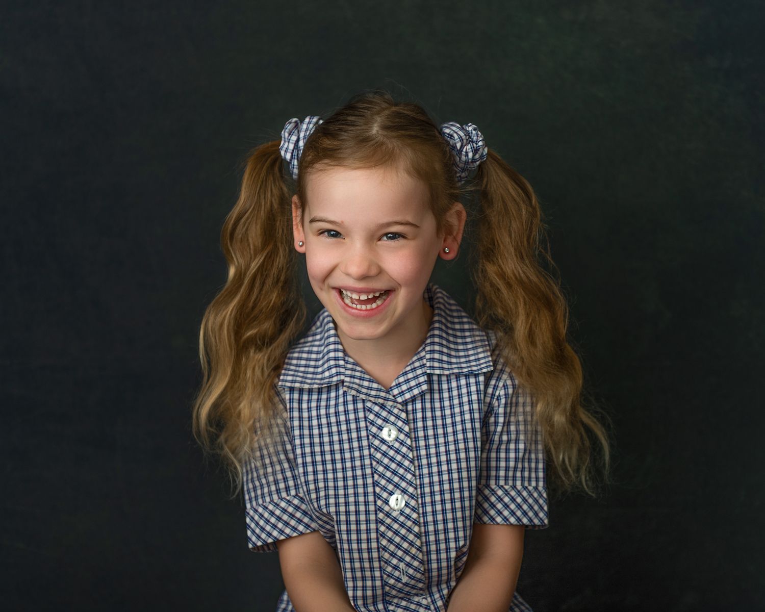 Child in gingham dress poses playfully against dark backdrop.