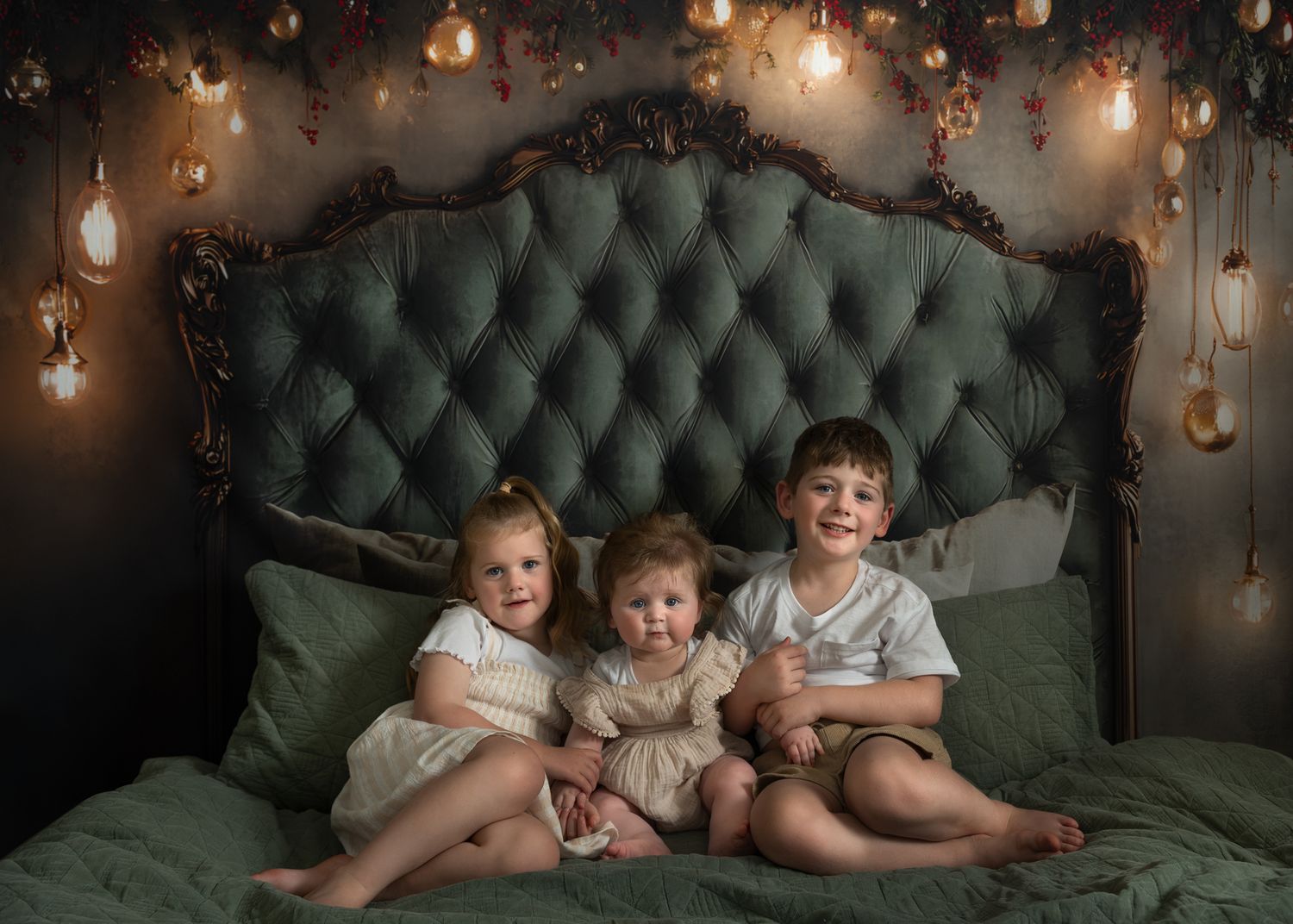 Three children sit on a green tufted headboard with hanging lights and greenery decorations above.