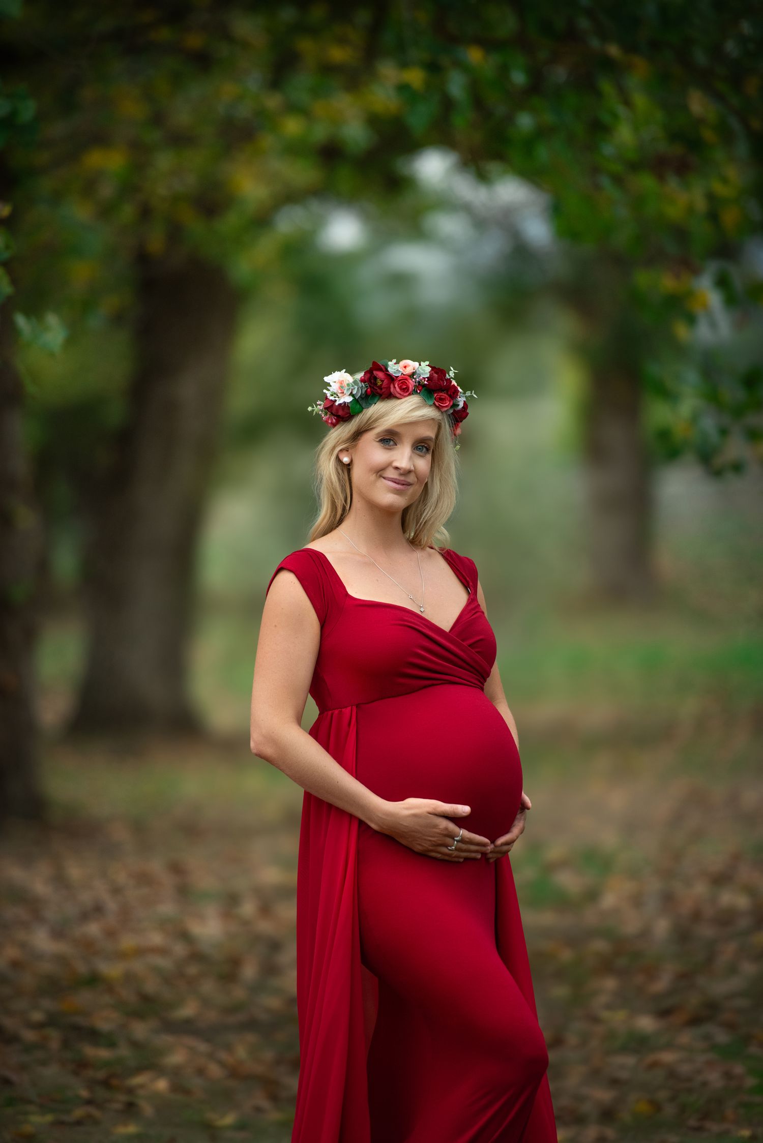 Maternity photo in red dress with floral crown standing among autumn trees in outdoor setting.