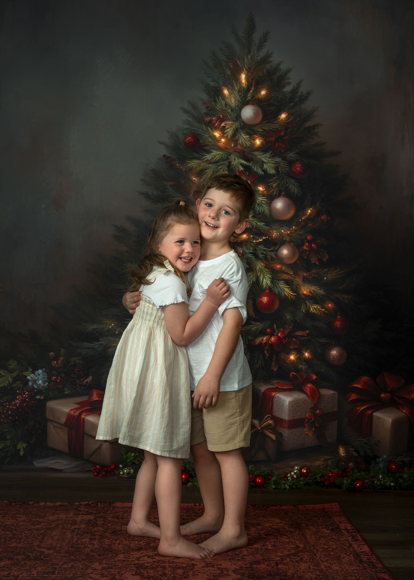Two children share a tender moment in front of a decorated Christmas tree with wrapped presents underneath.