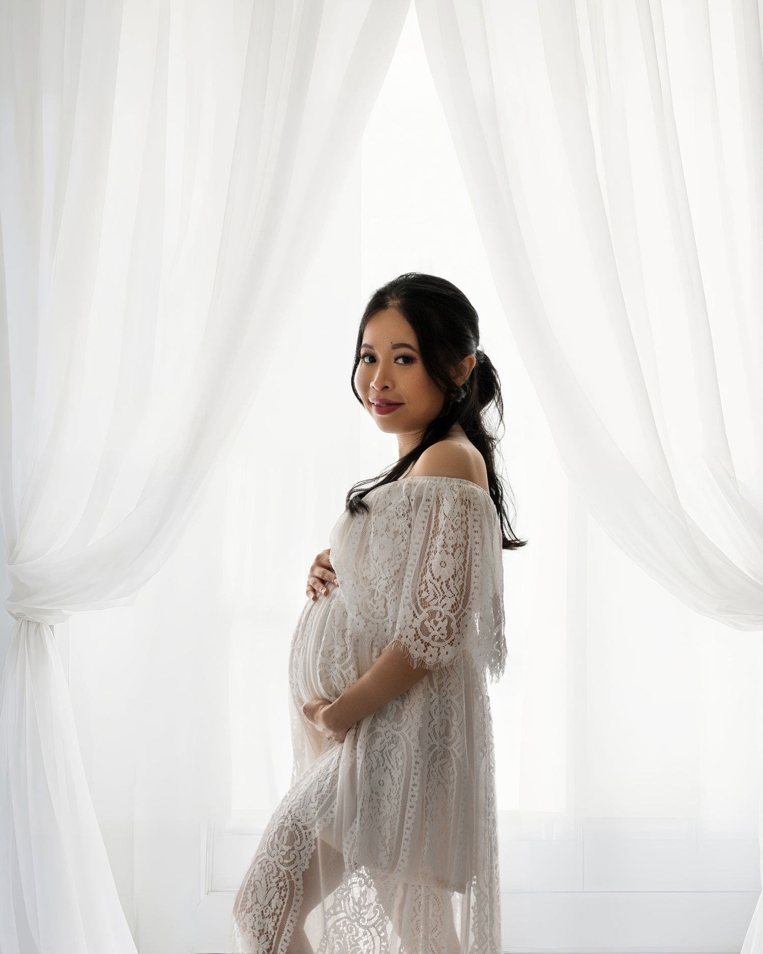Delicate white lace maternity gown photographed against bright white curtain backdrop.