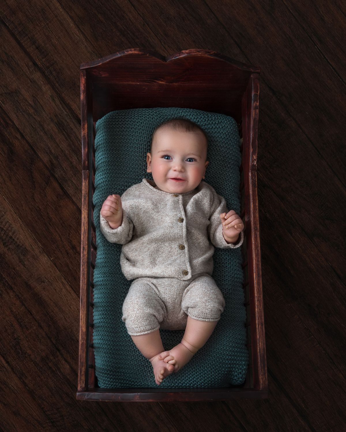 Baby in gray romper photographed from above in wooden box with teal cushion.