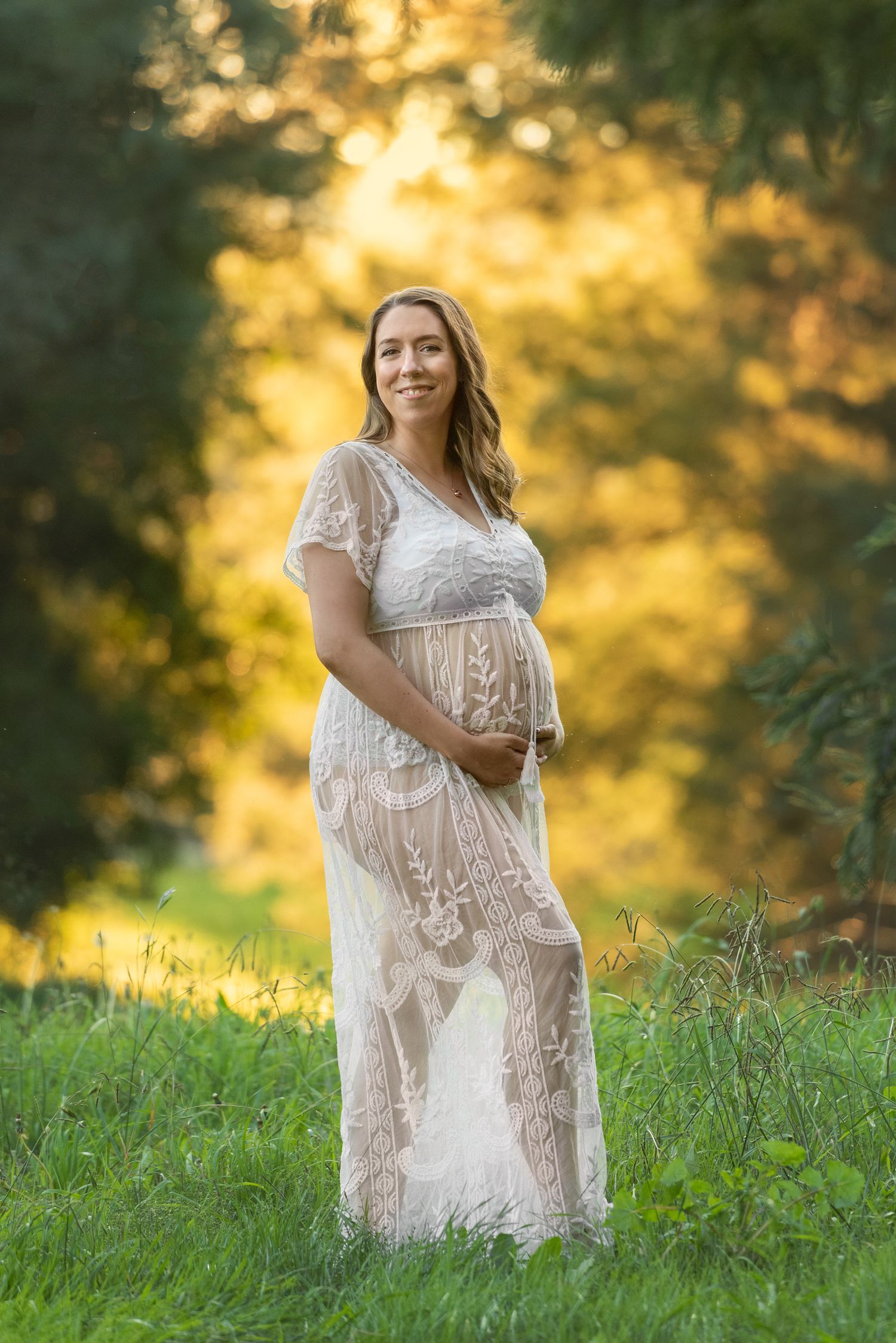 Maternity portrait in white lace dress against golden sunset in grassy field.