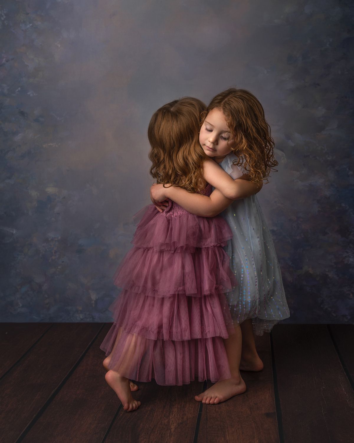 Two young children in tulle dresses share a sweet embrace against a dark backdrop.