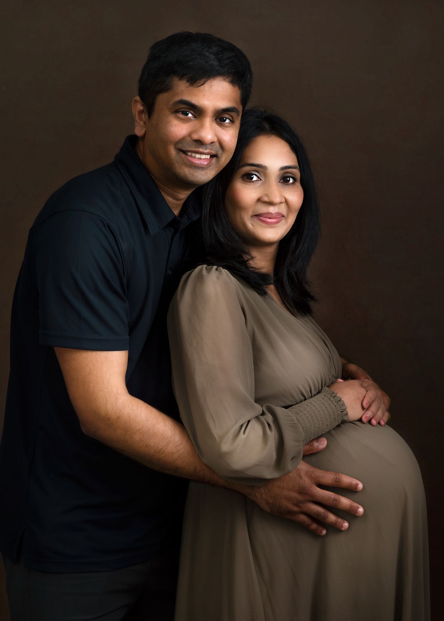 Studio maternity portrait with couple in casual attire against dark background.