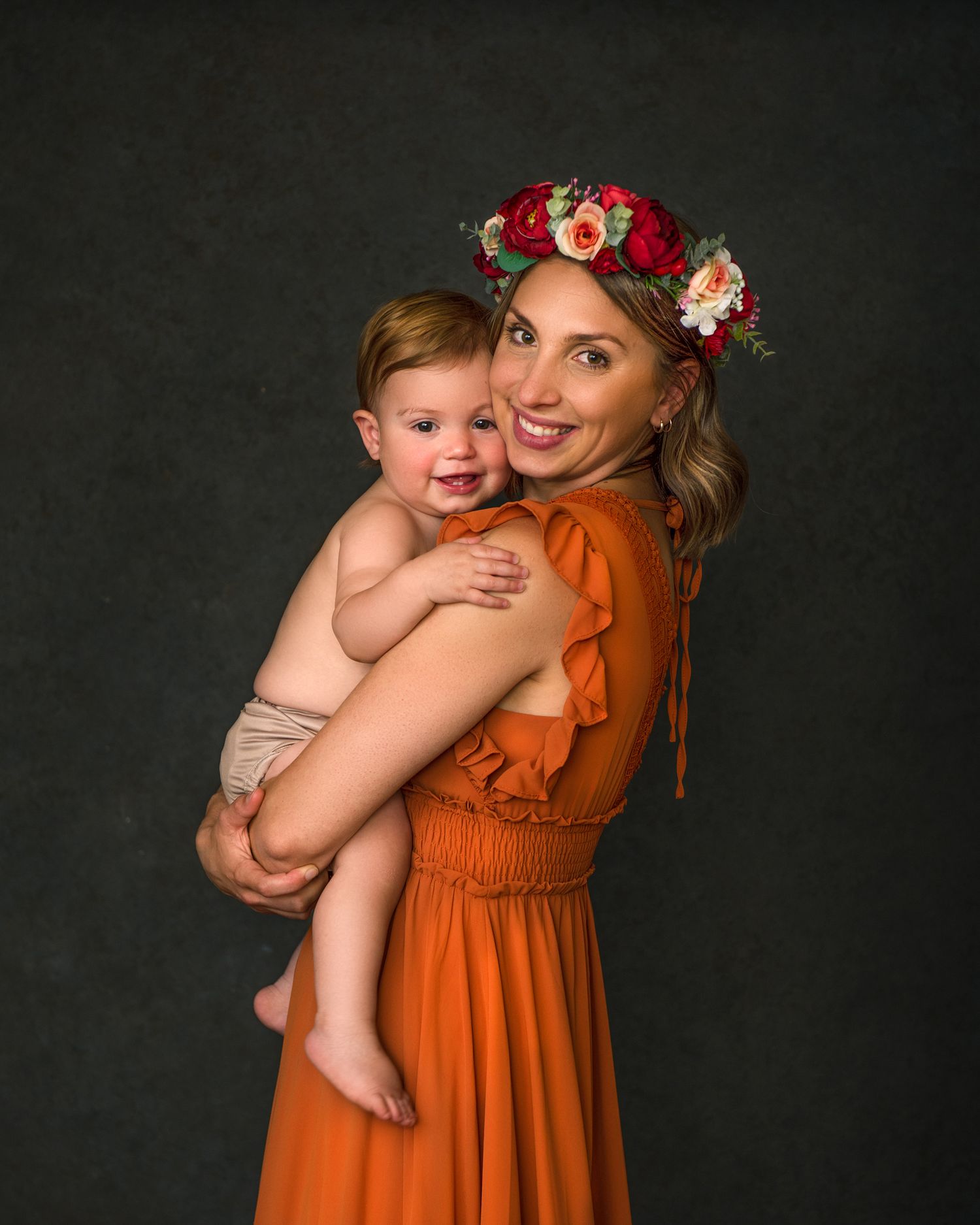 Mother in orange dress with floral crown holds smiling baby against dark backdrop.