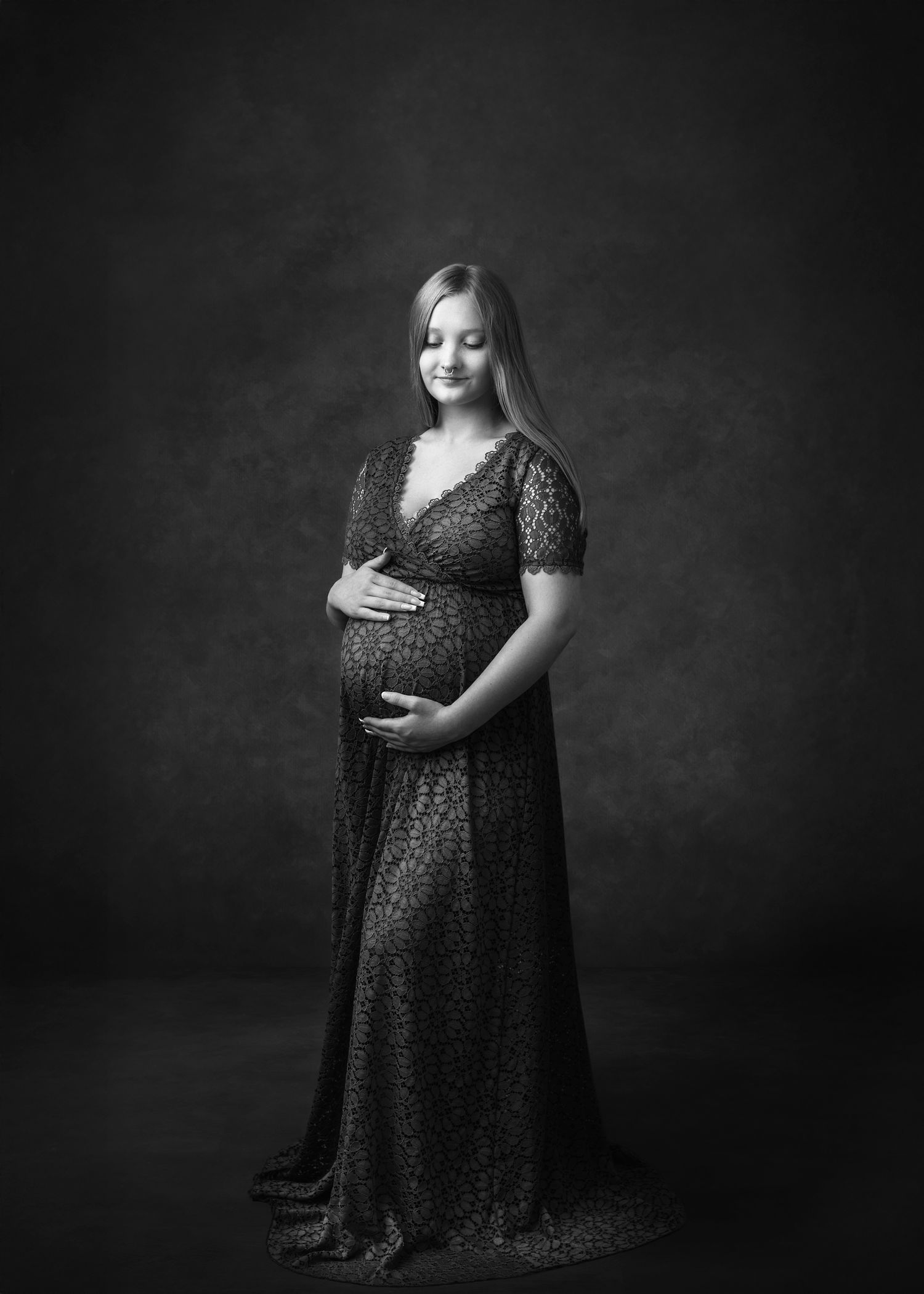 Studio maternity portrait in black lace dress against dark textured background.