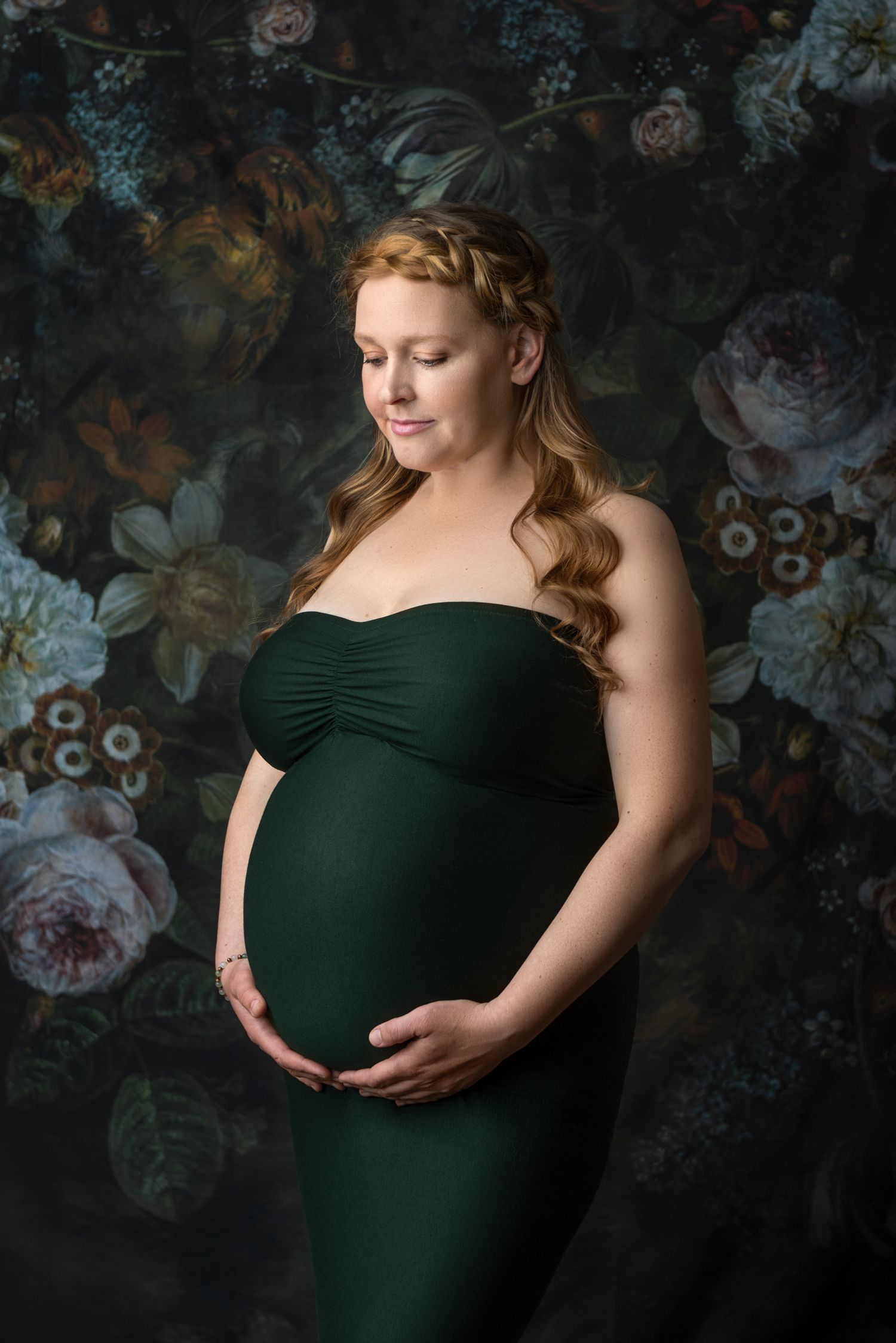 Maternity portrait in emerald green dress against floral backdrop with vintage styling.