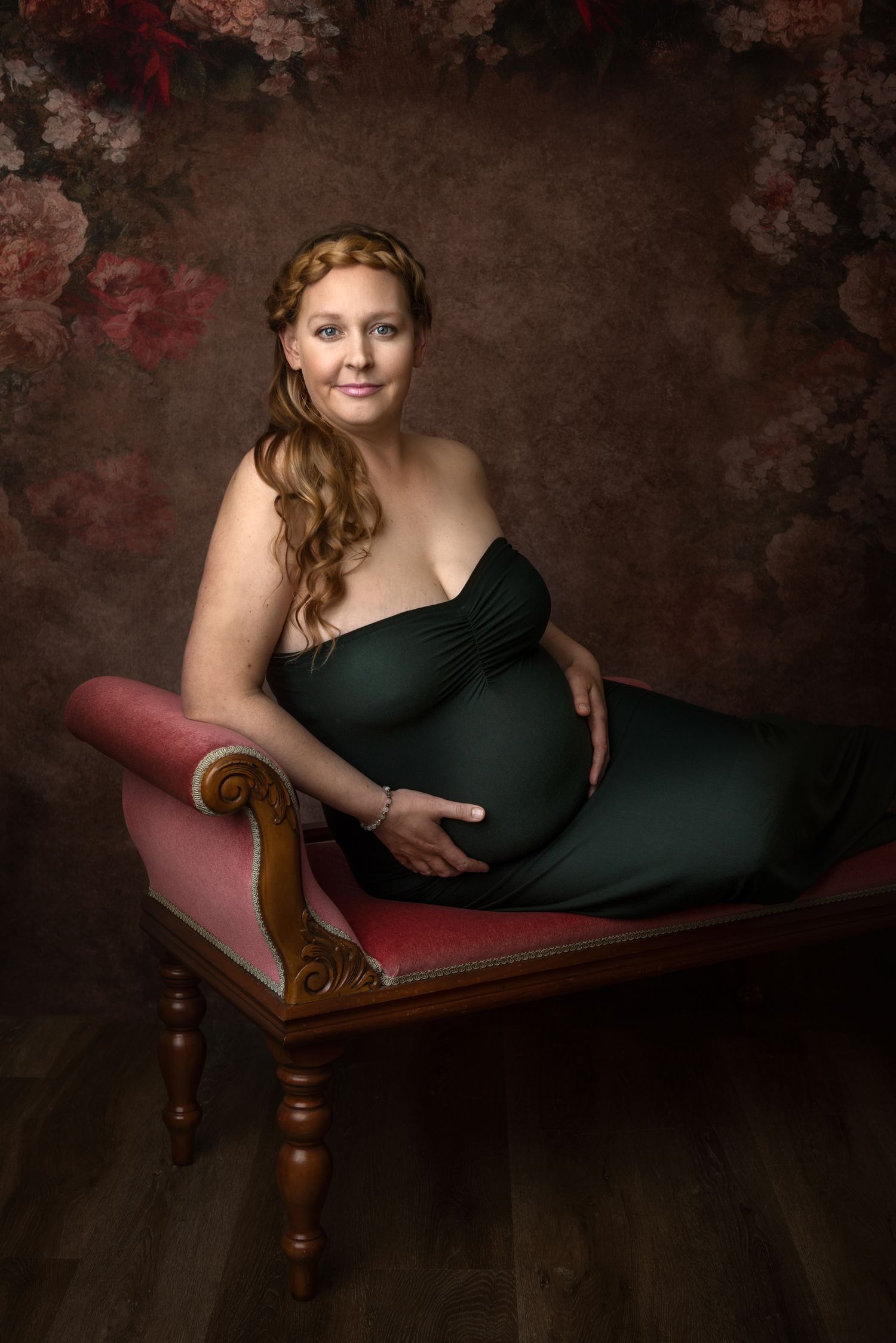 Elegant maternity session on vintage chaise lounge with dark floral background.