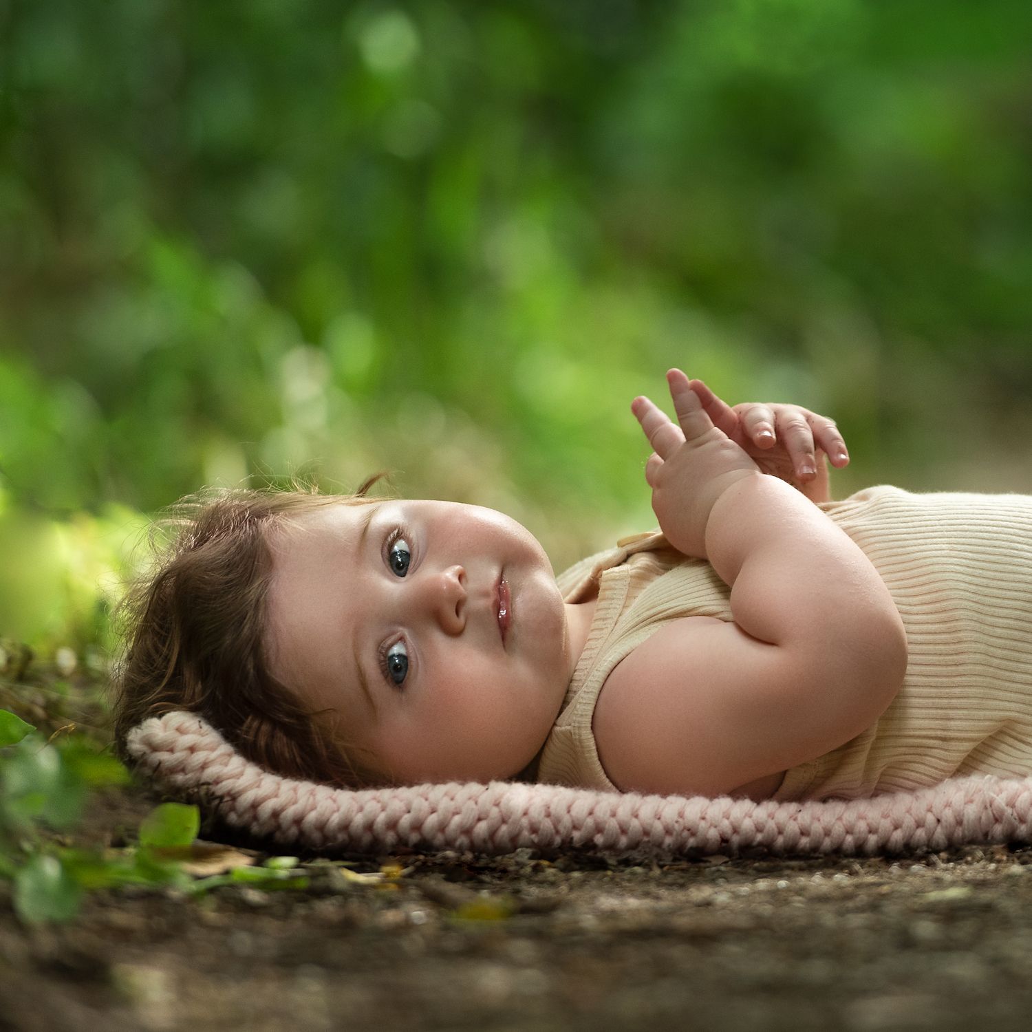 A baby in cream colored outfit lies on textured blanket outdoors in nature.