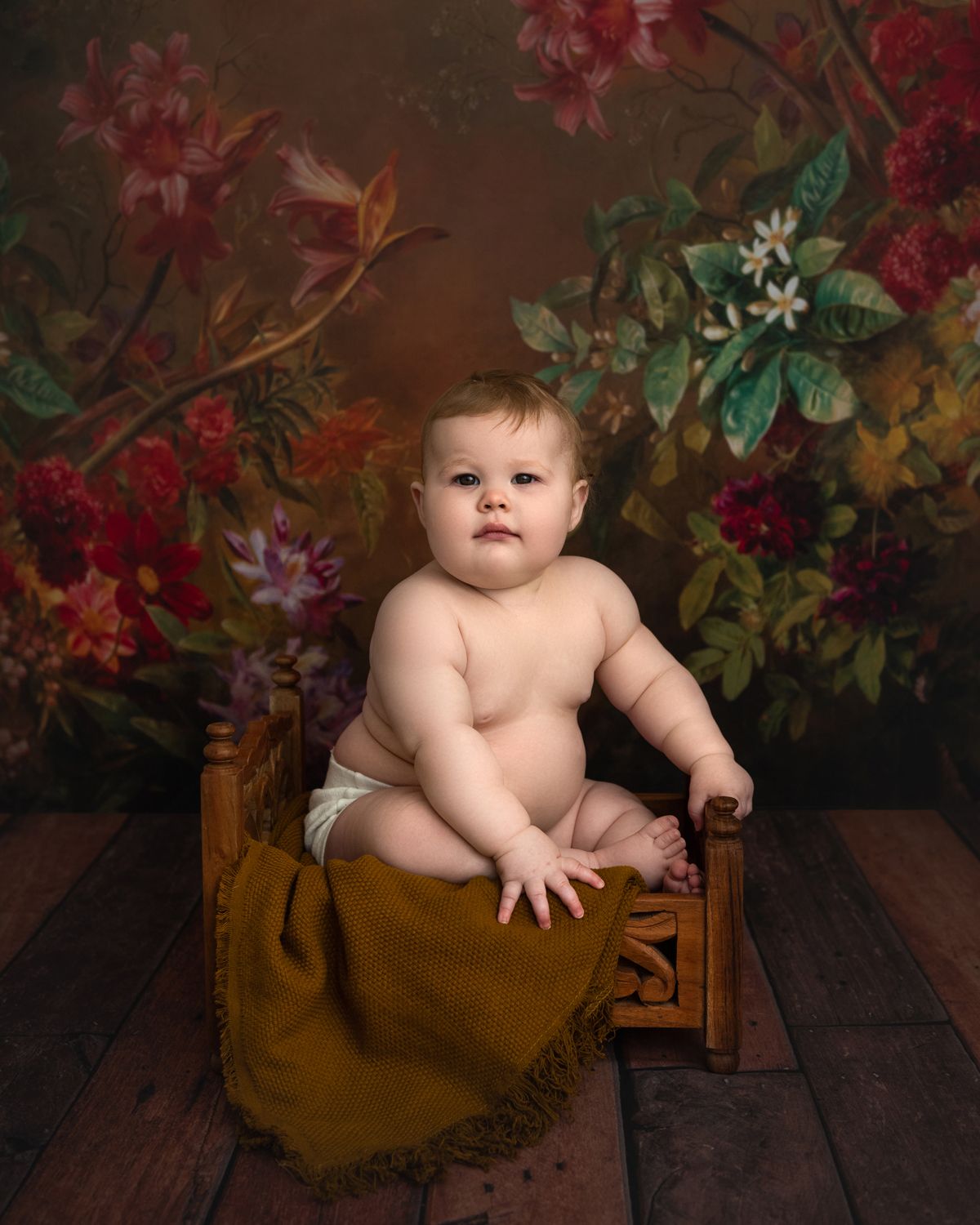 Baby sits in wooden crate surrounded by floral backdrop during studio photo session.