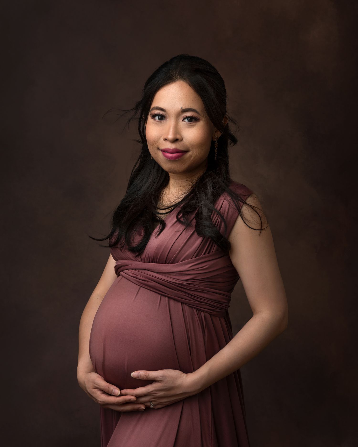 Studio maternity portrait in mauve wrap dress against dark background.