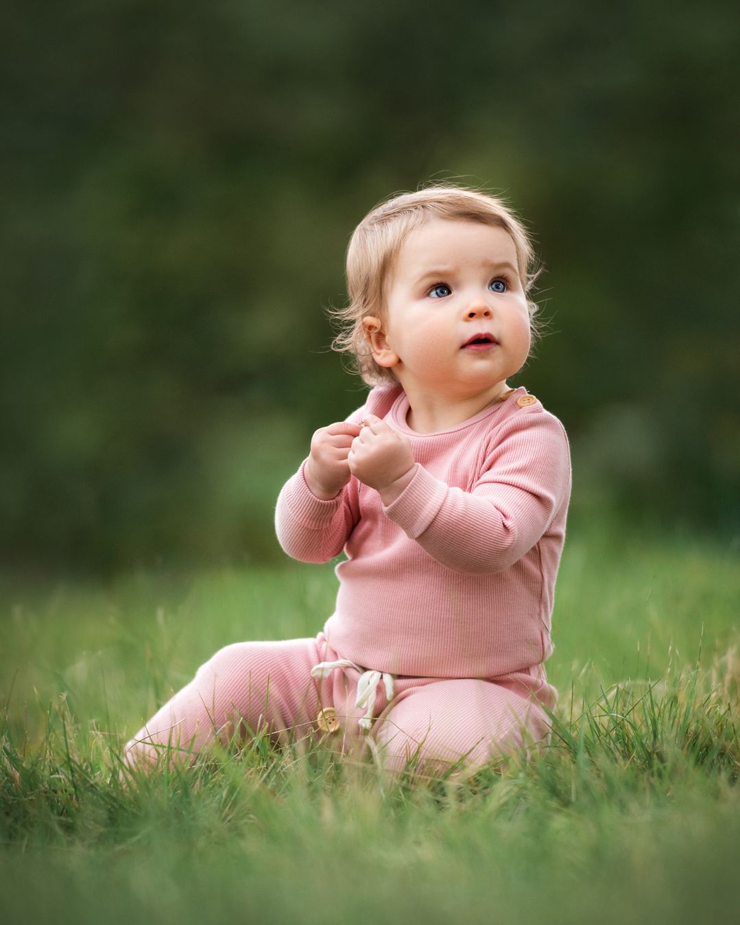 A baby in pink clothing sits in green grass during an outdoor photo session.