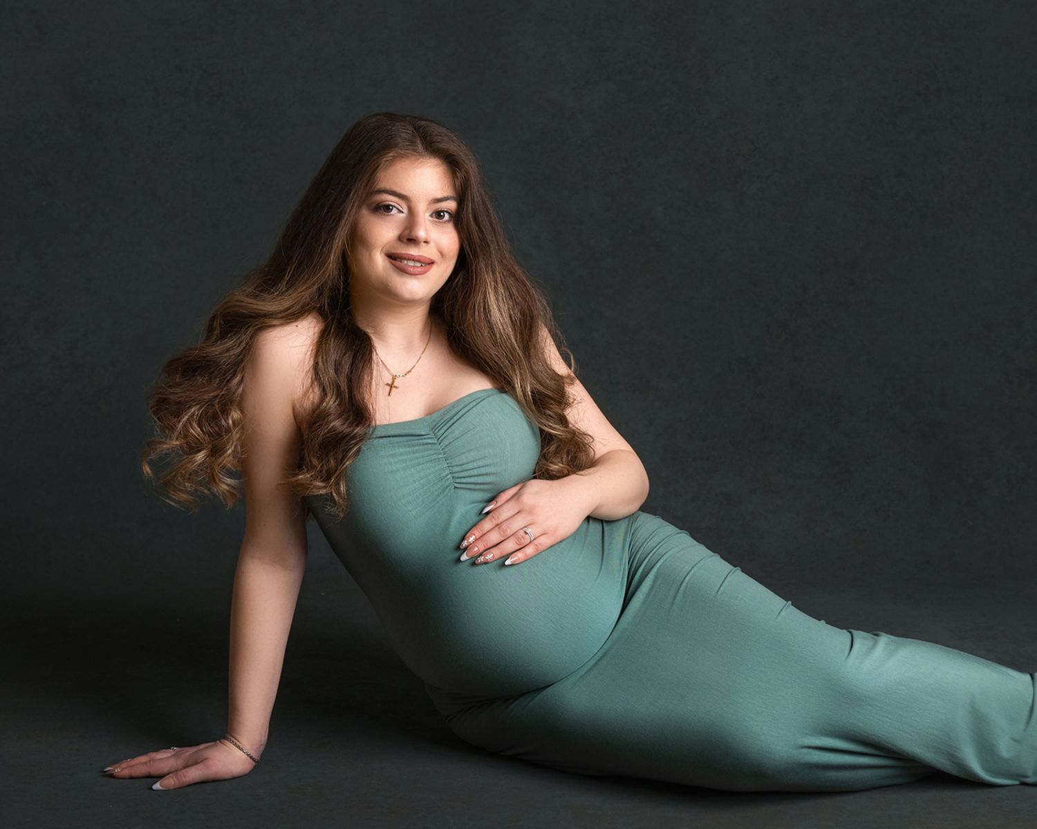 Woman in teal dress poses gracefully on dark background in artistic maternity photo series.