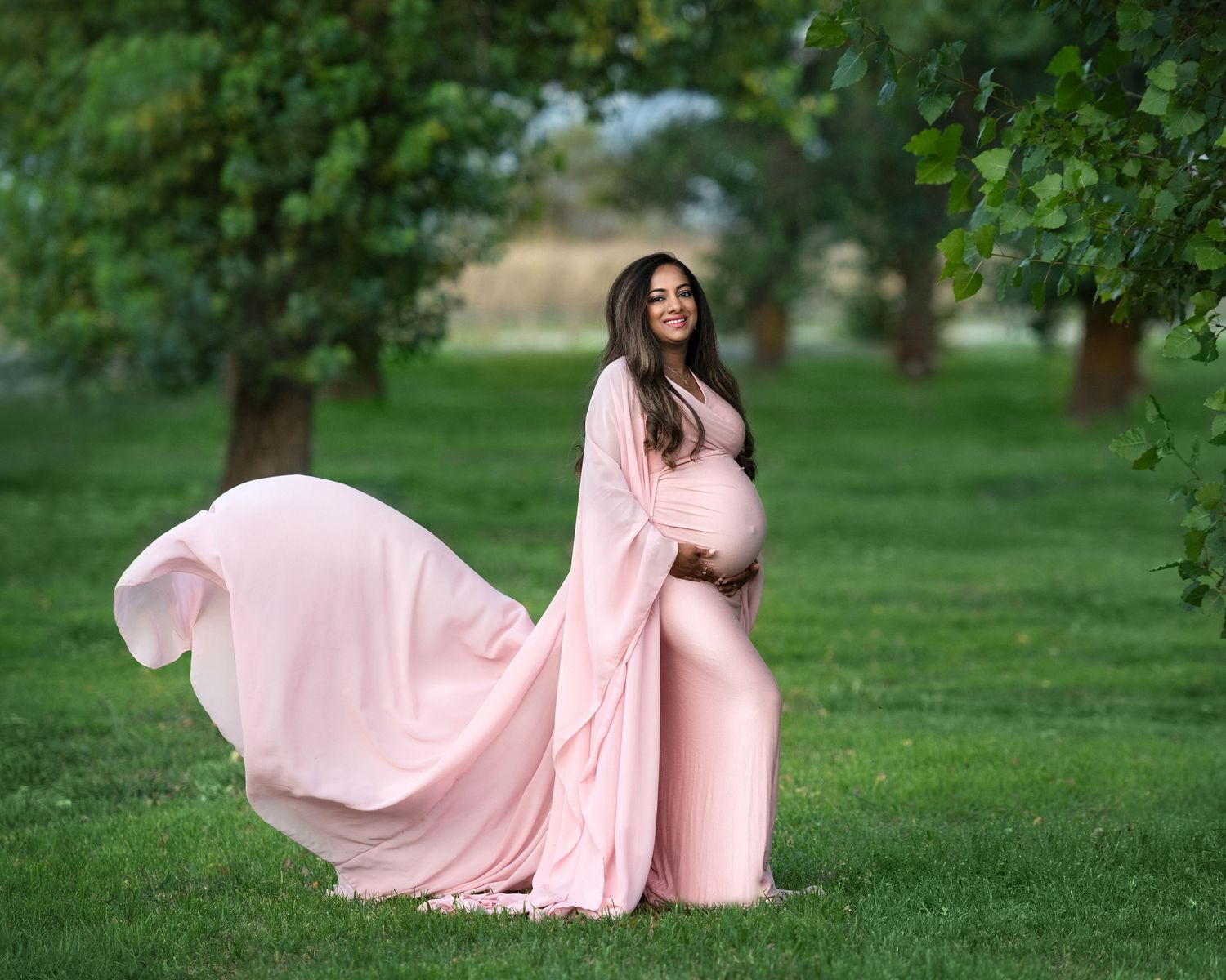 Flowing pink maternity gown billows in breeze during outdoor garden photoshoot.