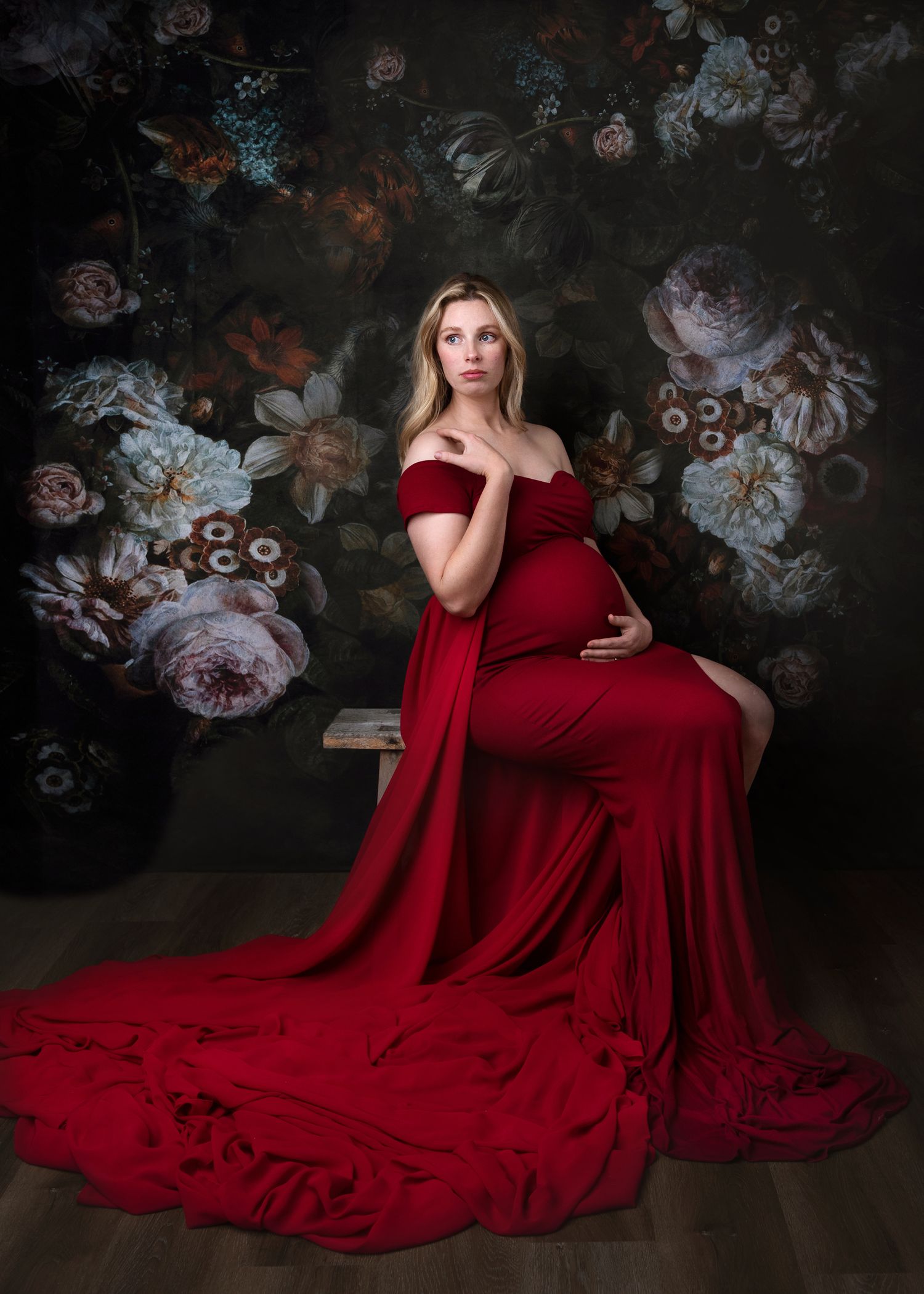 Deep red gown drapes elegantly against moody floral backdrop in artistic portrait session.