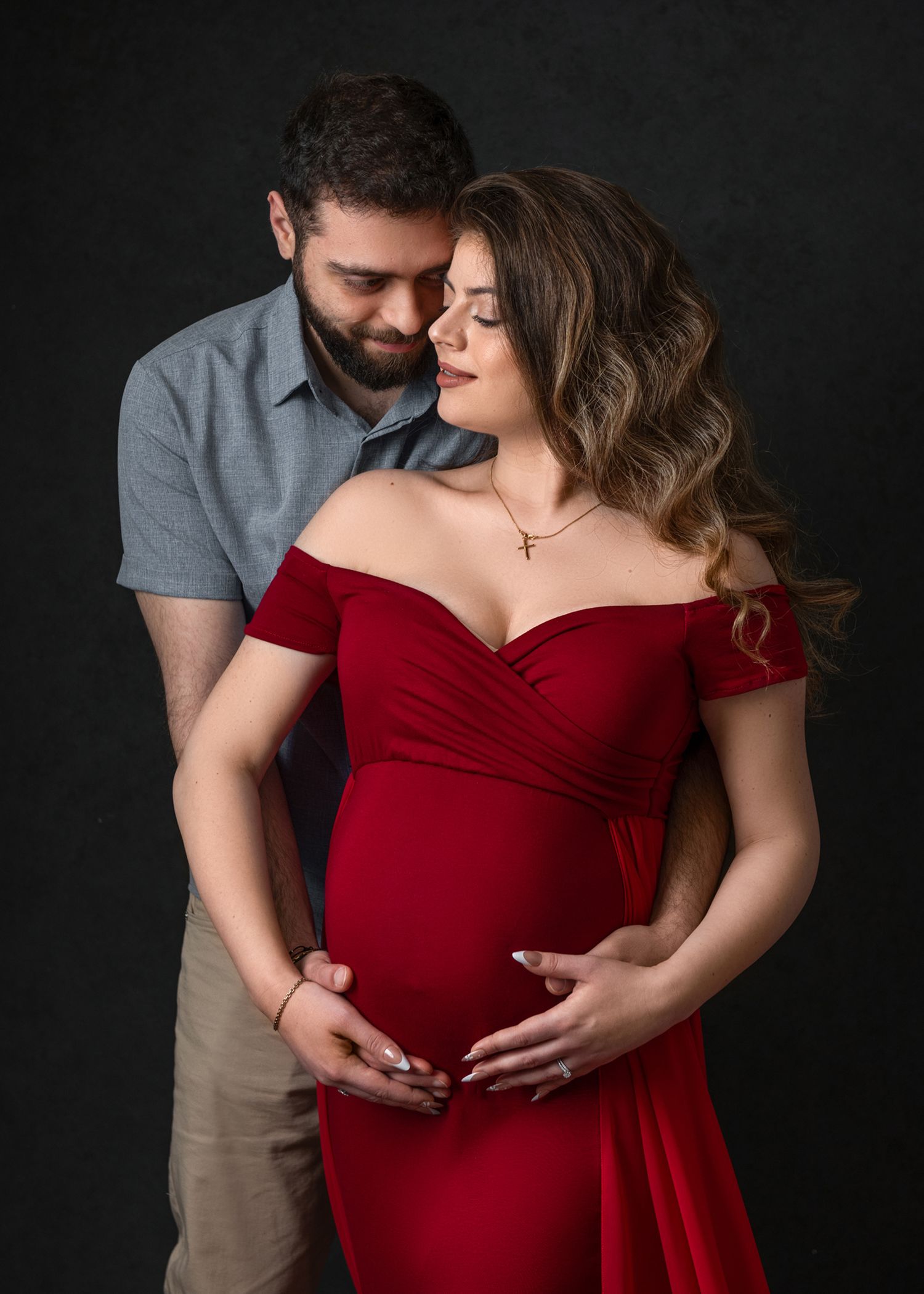 Intimate maternity portrait in red dress with couple embracing against dark background.