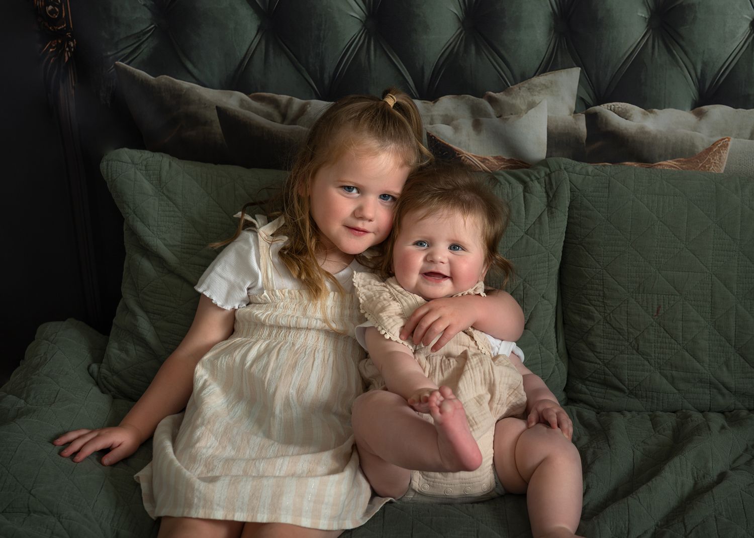 Two children in white outfits sit together on a green couch sharing a tender moment.