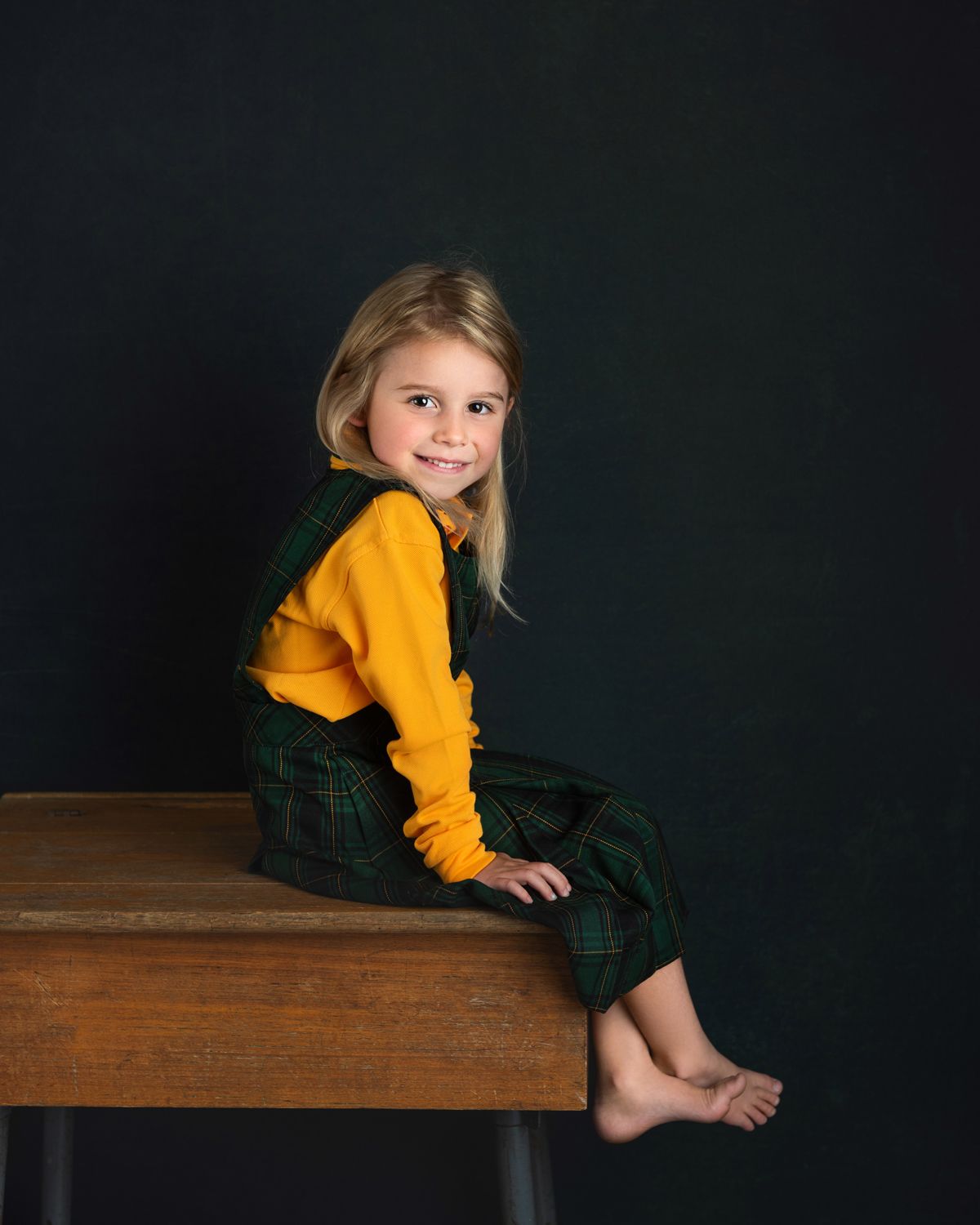 Child wearing a yellow sweater and plaid skirt sits on a dark wooden surface against black background.