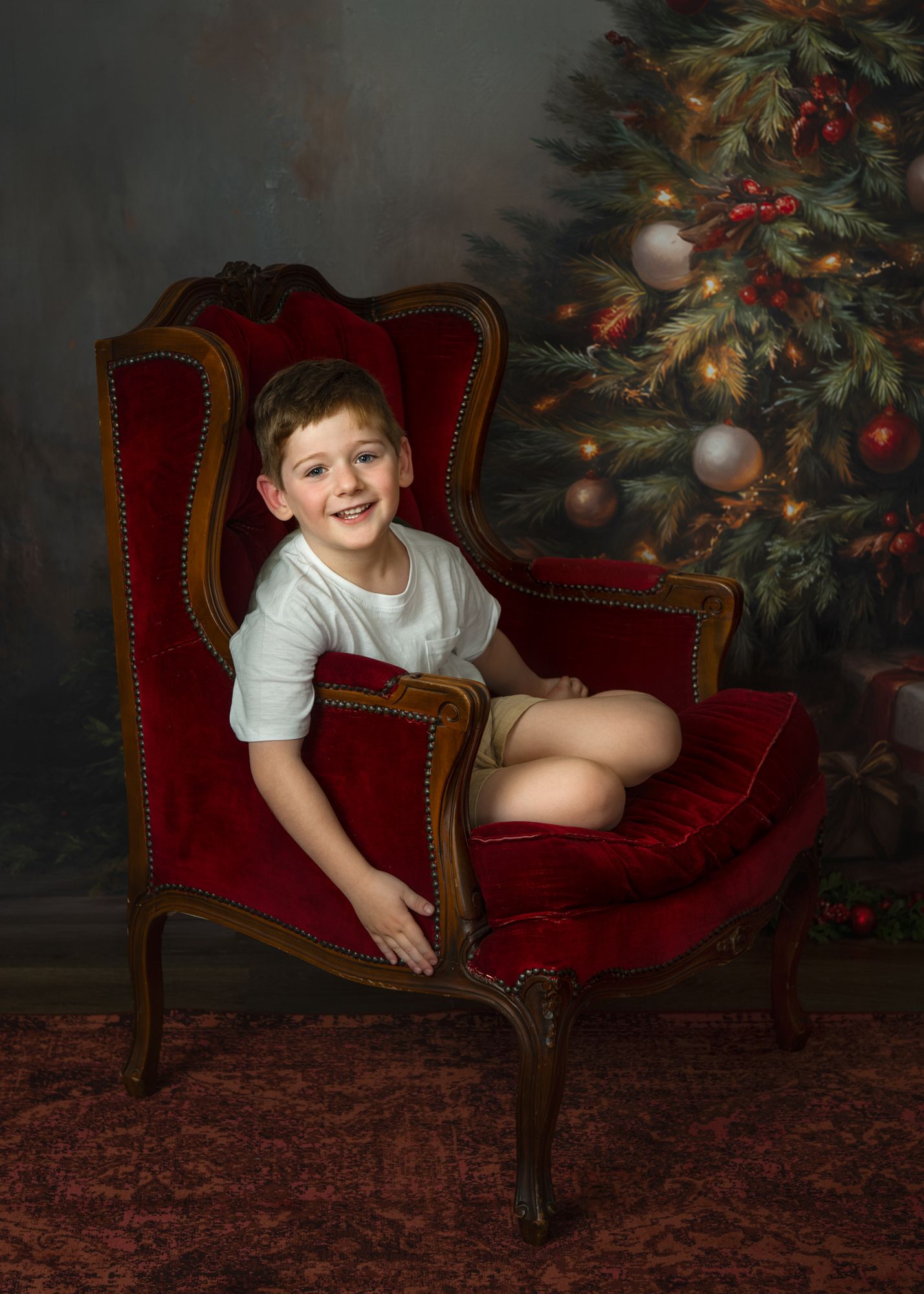 Child lounges playfully on red velvet chair next to decorated Christmas tree with twinkling lights.