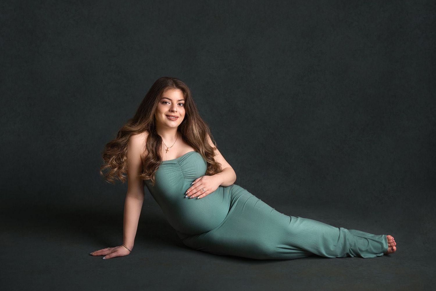 Artistic maternity photo in teal dress with flowing fabric against dark backdrop.