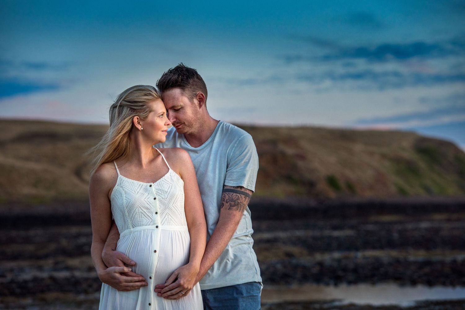 Romantic maternity photo of couple embracing on beach at sunset with mountains in background.