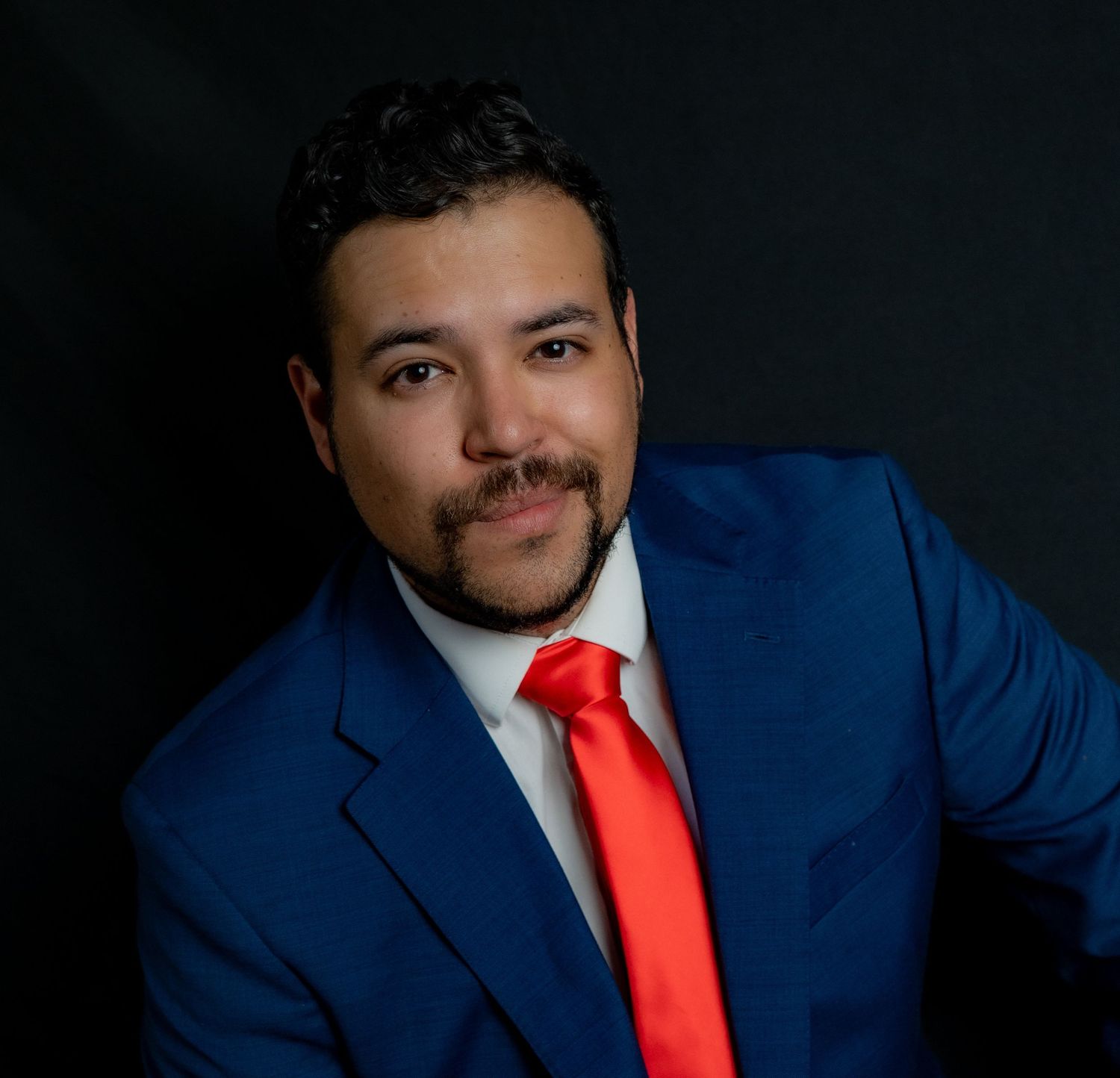 Professional headshot of someone in a royal blue suit with bright red tie against a dark background.