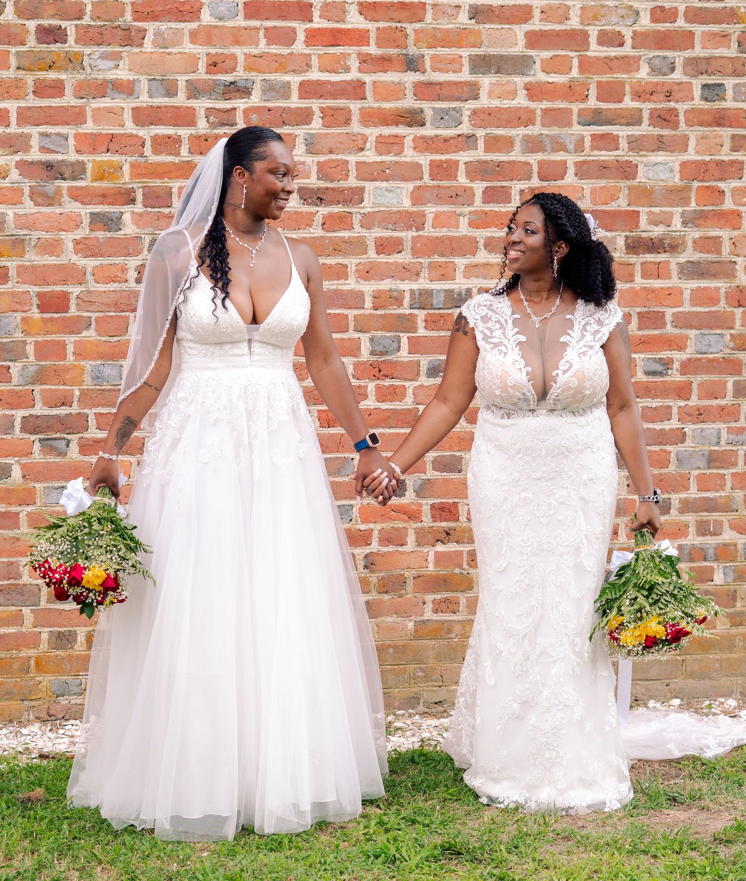 Two brides in flowing white gowns stand together against a rustic brick wall with bright bouquets.