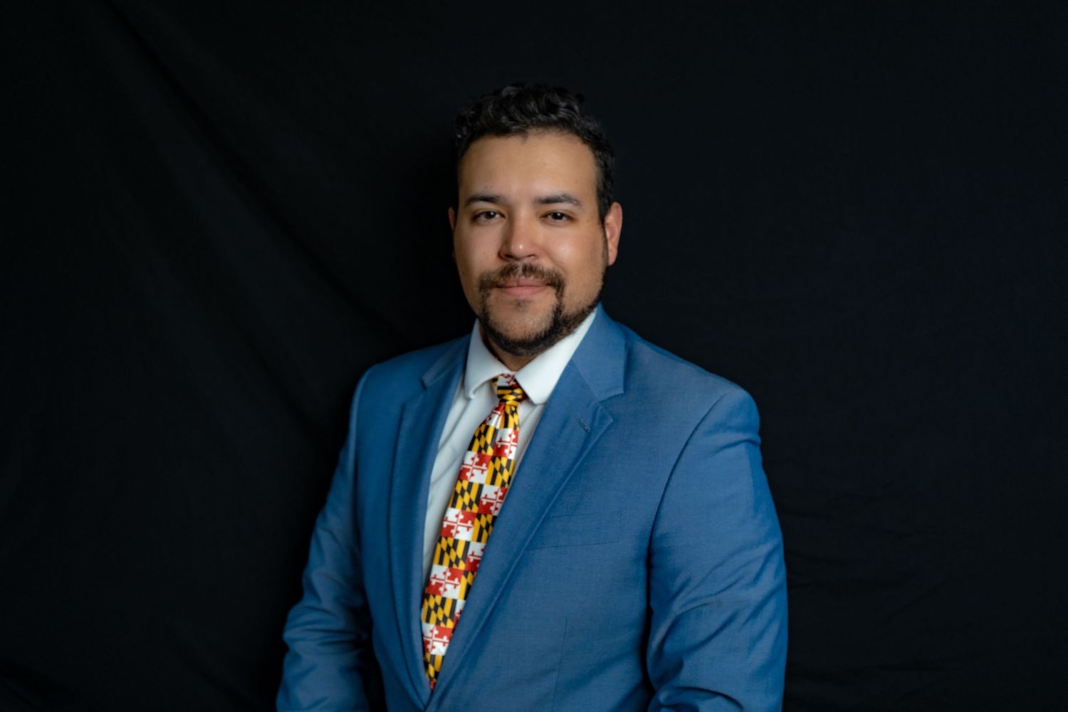 Professional portrait of someone in a bright blue suit jacket and colorful geometric patterned tie against a dark background.
