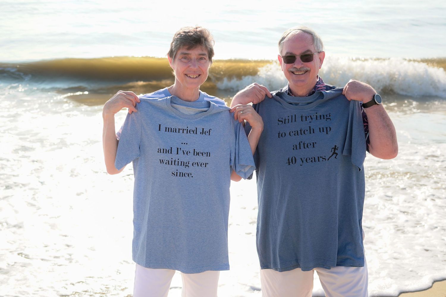Two people wearing matching t-shirts pose at a beach during sunset while smiling at the camera.