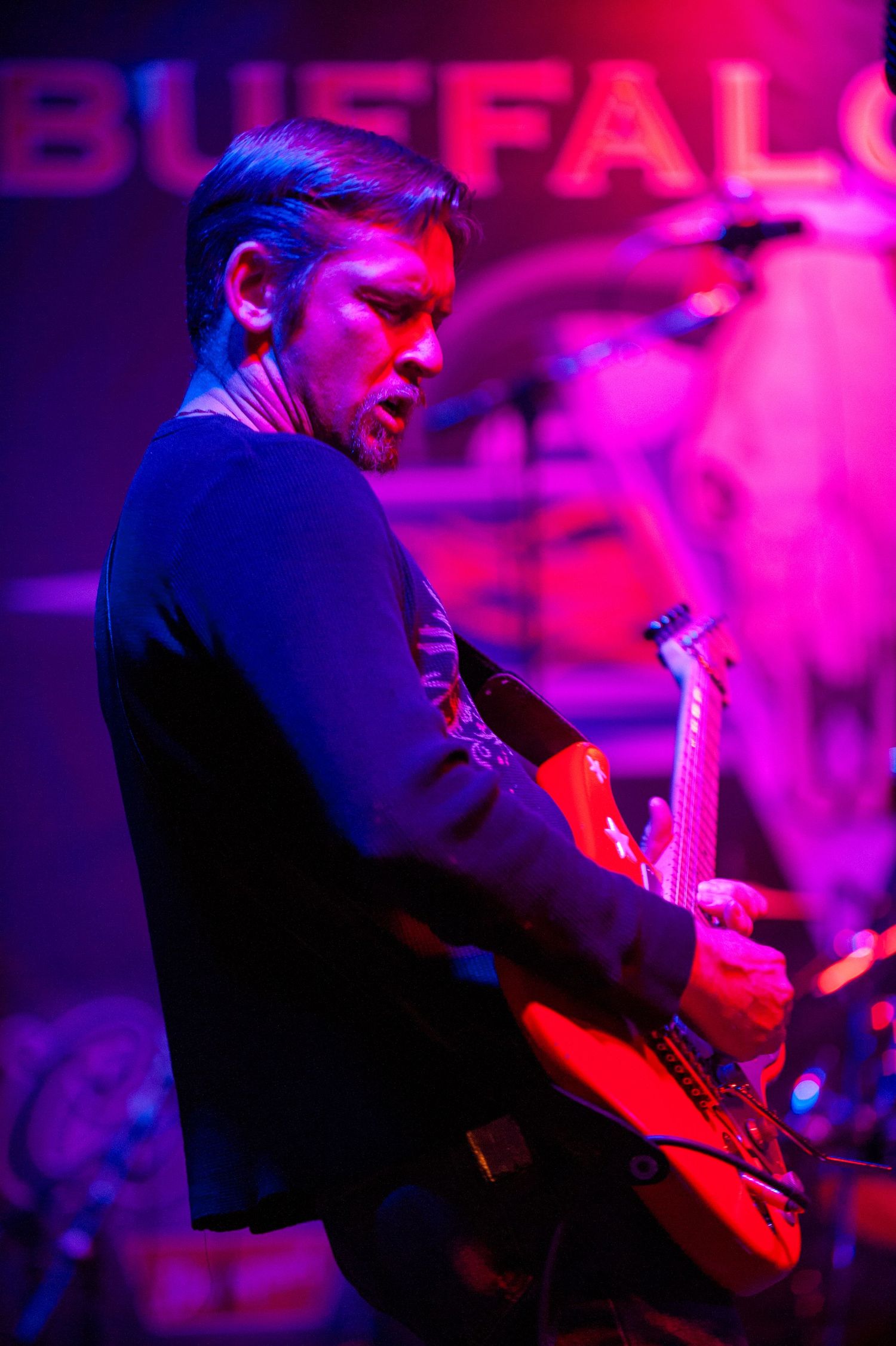 Guitarist performs on stage illuminated by dramatic purple and red lighting.