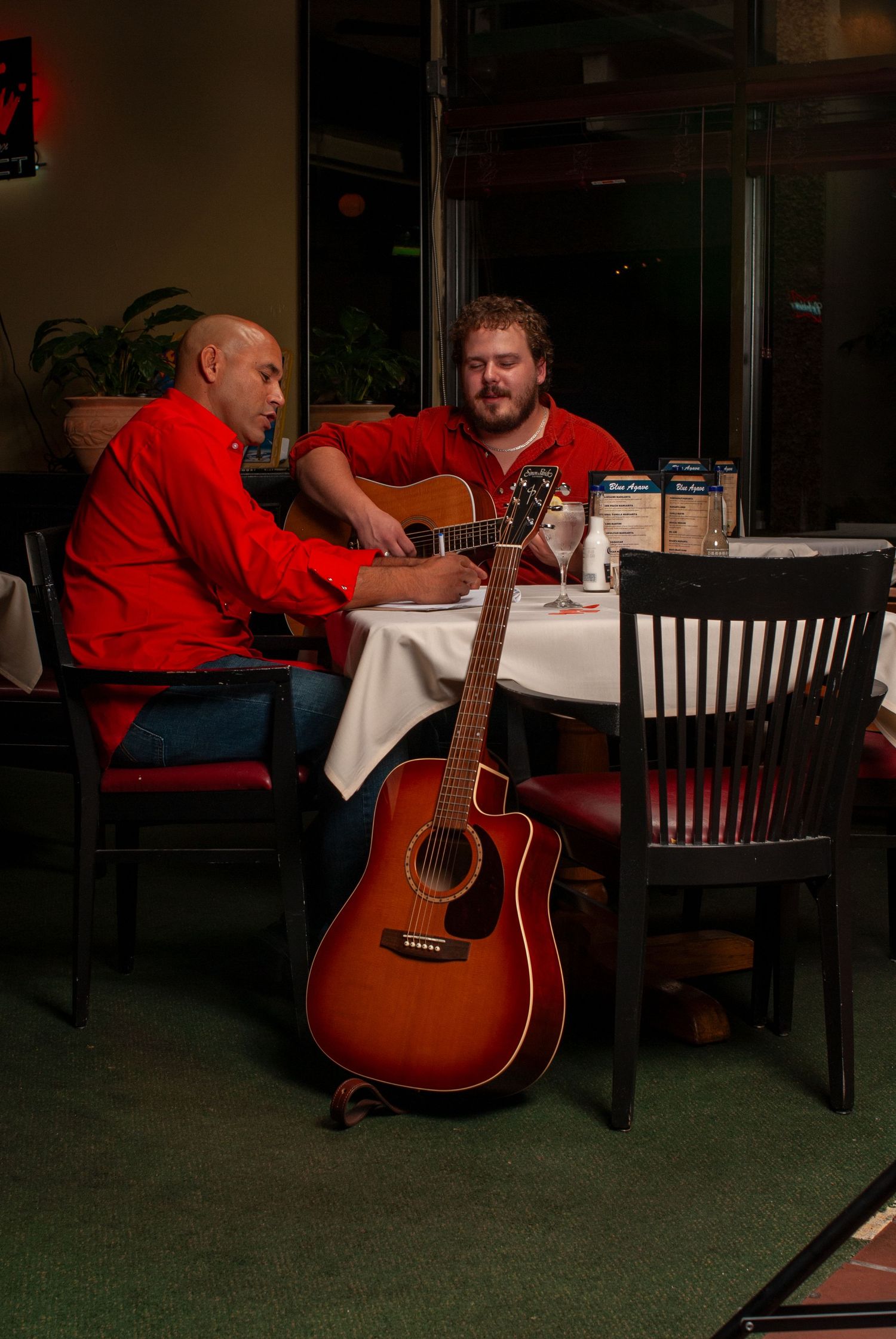 Musician playing acoustic guitar in a casual restaurant setting with green carpeting.