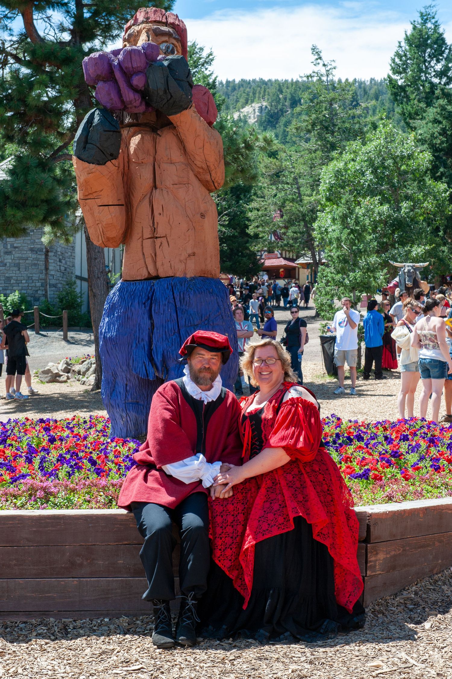 People in period costumes rest on a bench surrounded by vibrant flowers at an outdoor festival.