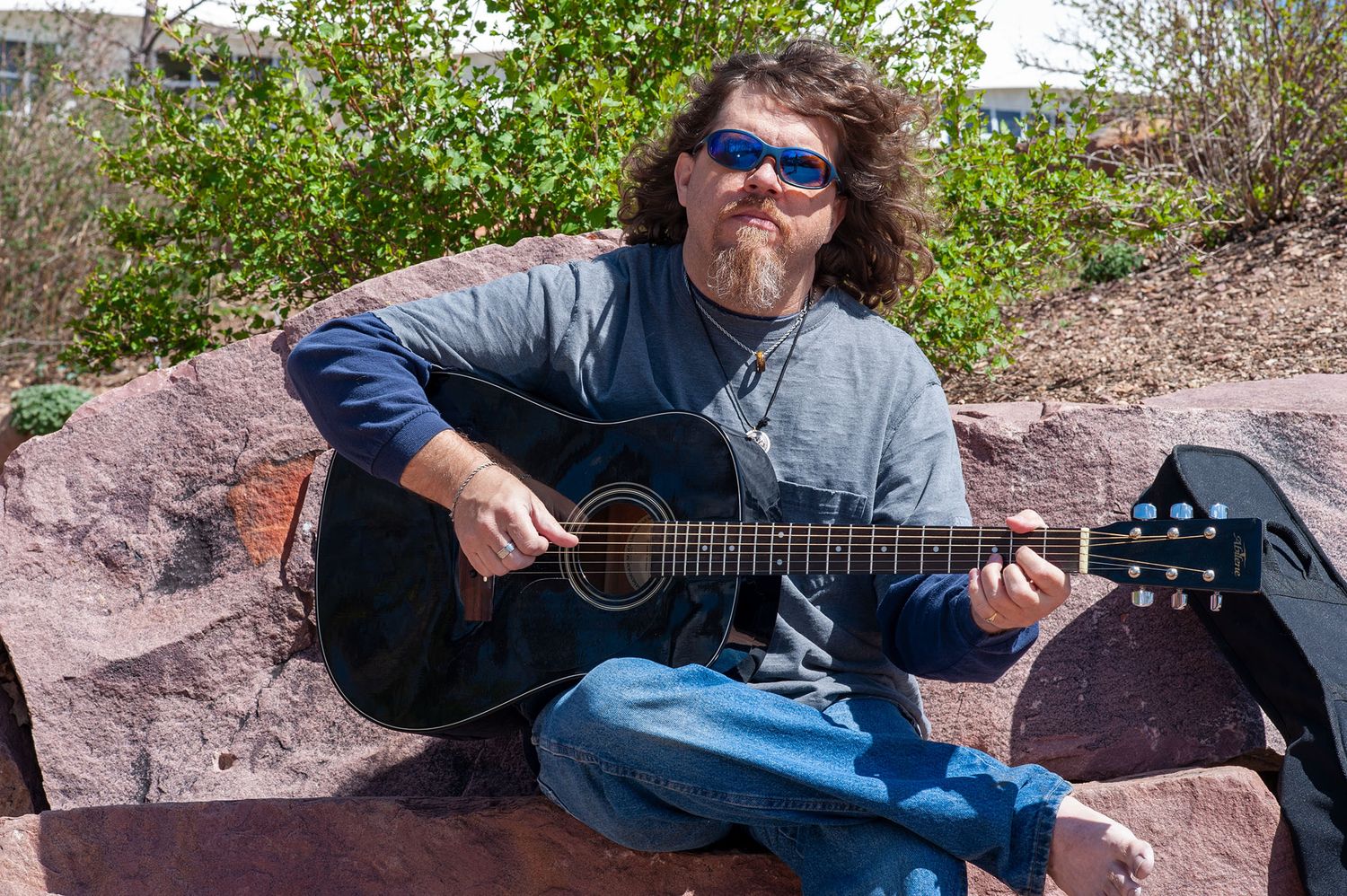 Person with sunglasses playing guitar while sitting on rocky terrain outdoors.
