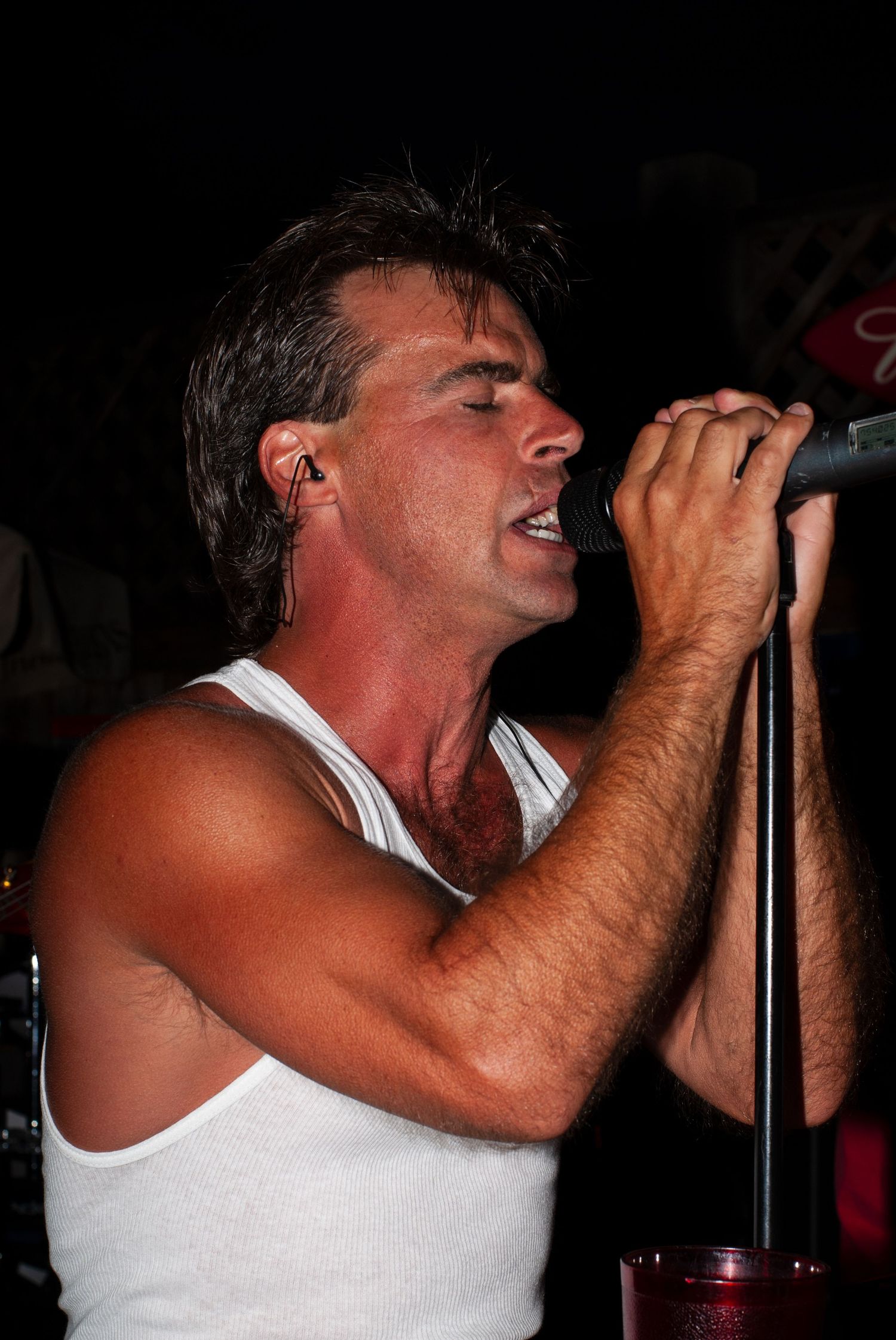 Singer performs passionately into microphone on dark stage in white tank top.