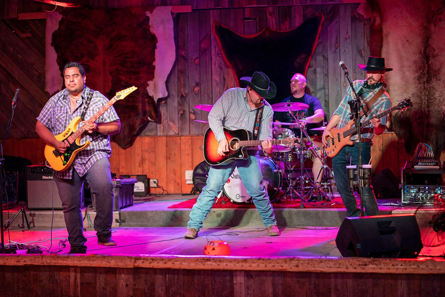 Country band performs live on rustic wooden stage with guitars and drums under colorful stage lighting.