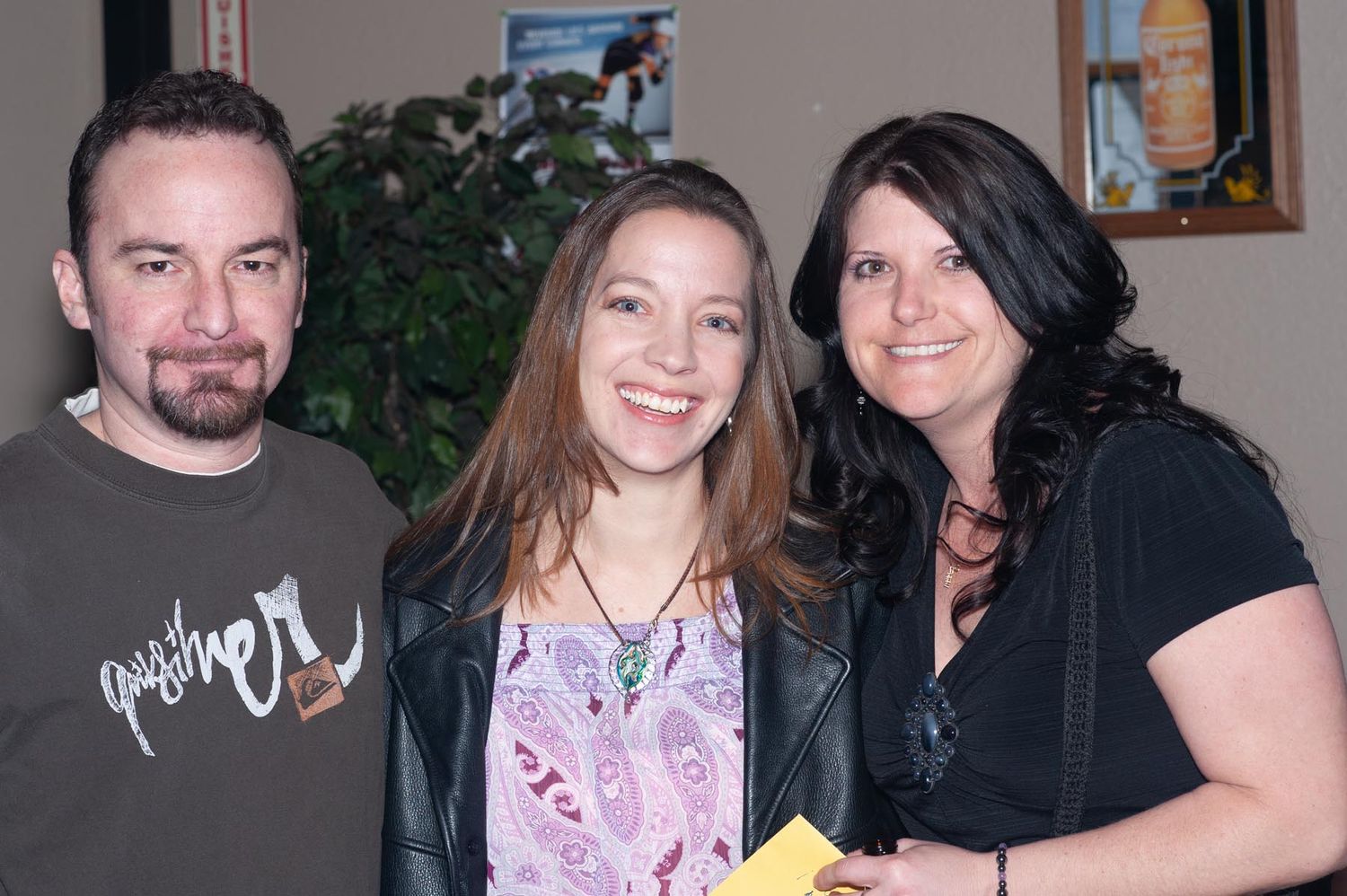 Three people stand together smiling at a casual indoor gathering with a house plant visible in the background.