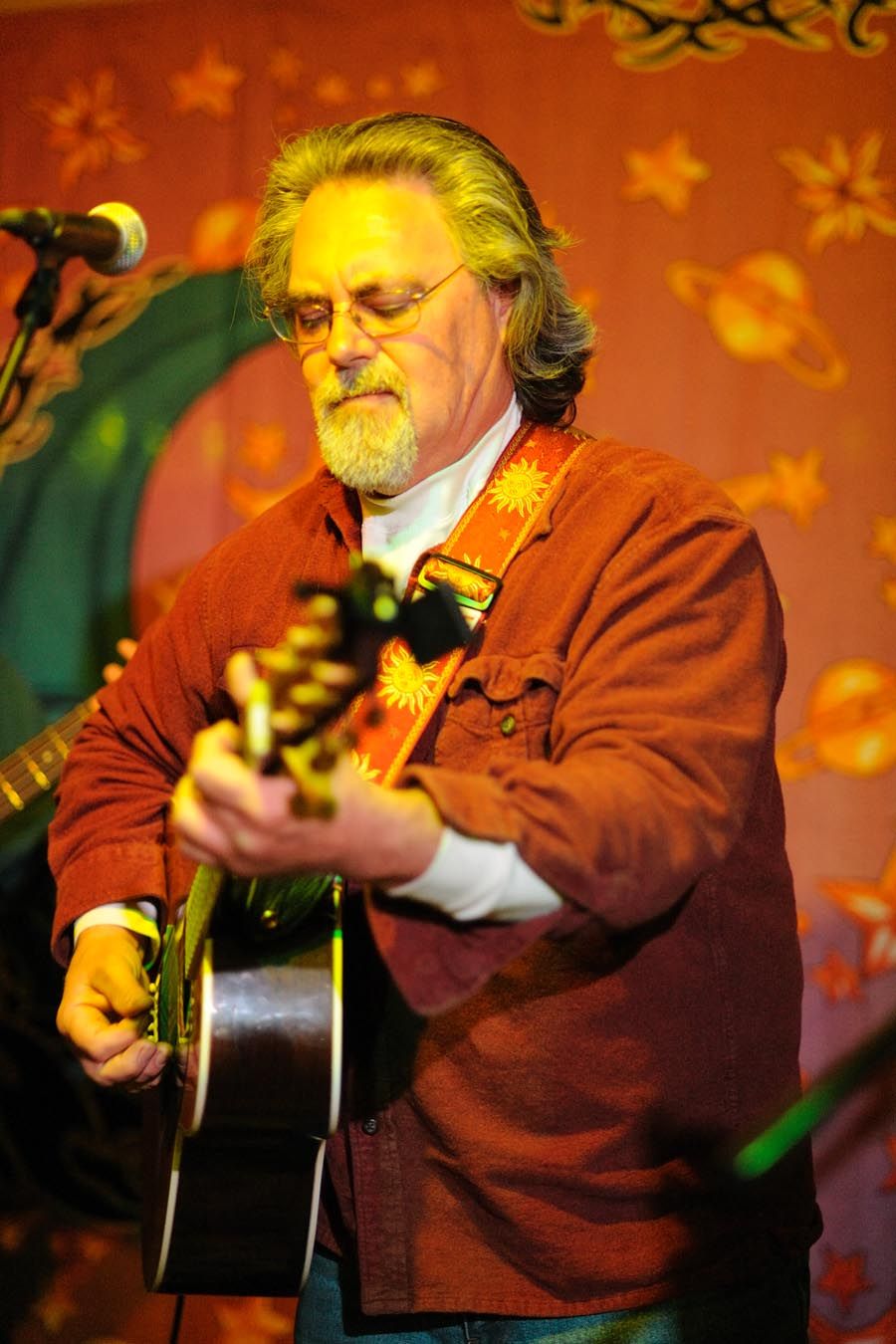 Musician in a brown sweater plays acoustic guitar against an orange backdrop with star decorations.
