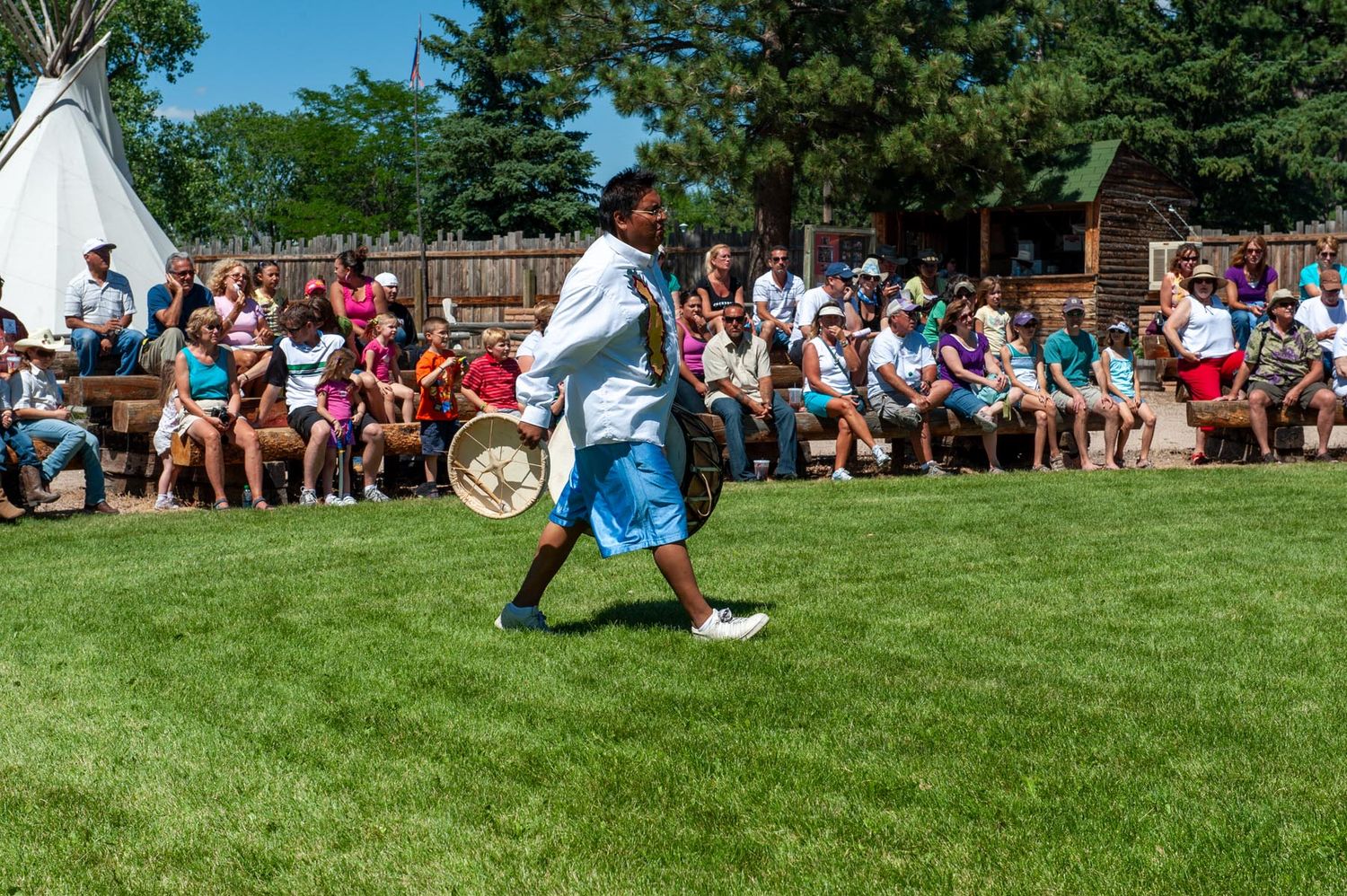 A crowd of spectators watches a sporting event on a grassy field surrounded by trees on a sunny day.