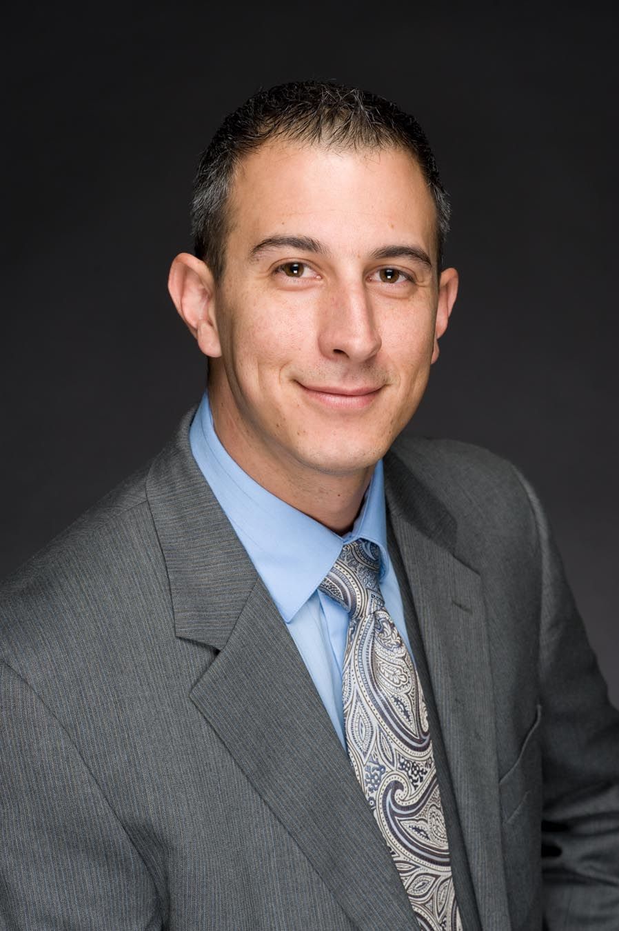 Professional headshot of someone in grey suit and patterned tie against dark background.