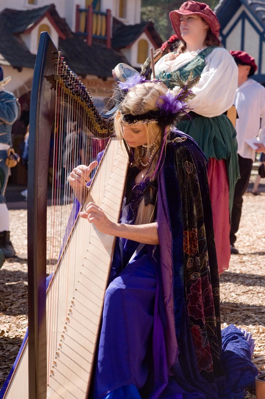 Musicians in renaissance fair costumes perform with harps outdoors.